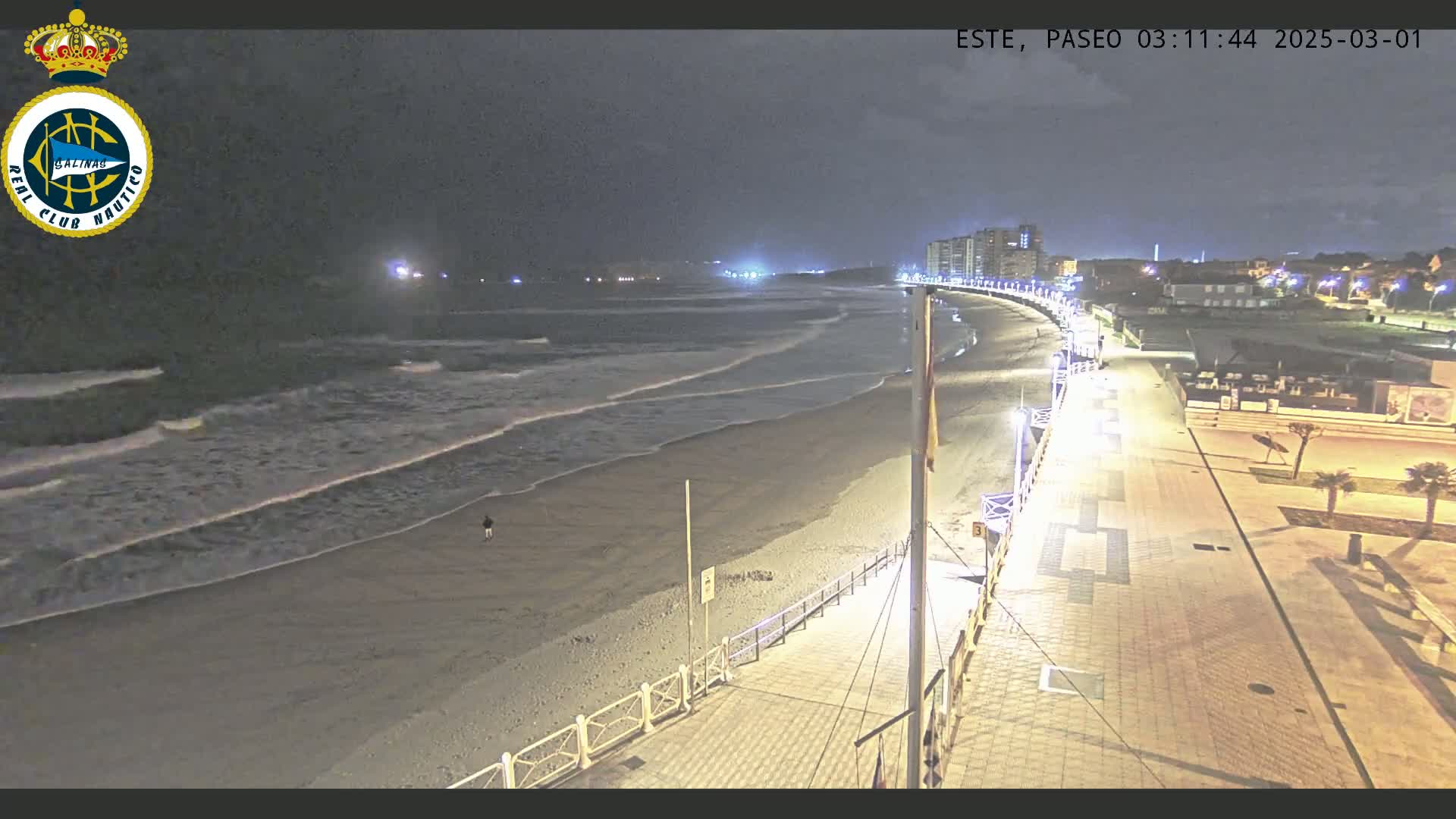 A nighttime beach scene shows gentle waves lapping the shore, a mostly empty beach, and a well-lit boardwalk curving along the coast.