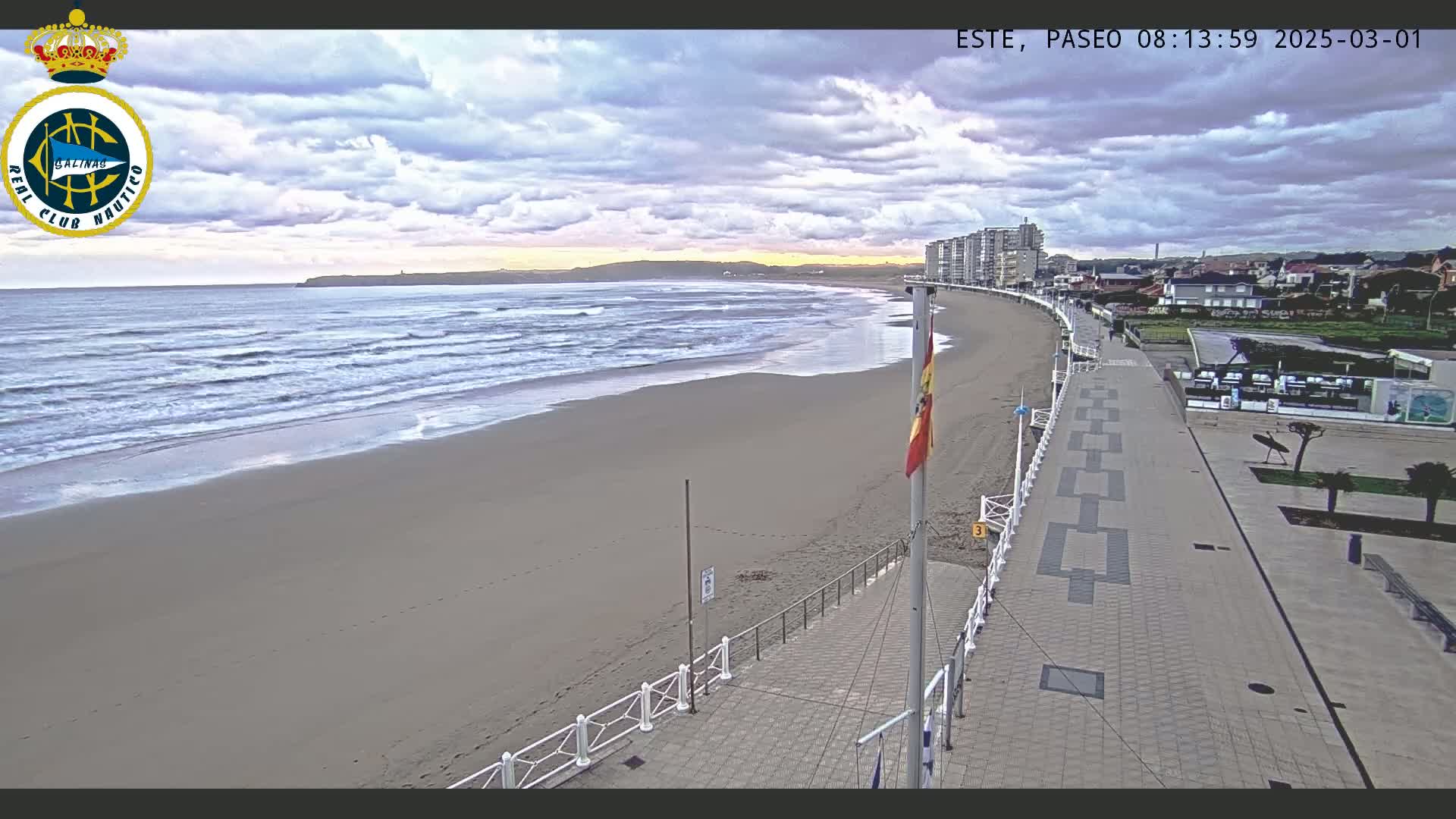 A sandy beach with gently rolling waves under a cloudy sky is bordered by a paved walkway leading to buildings in the distance.