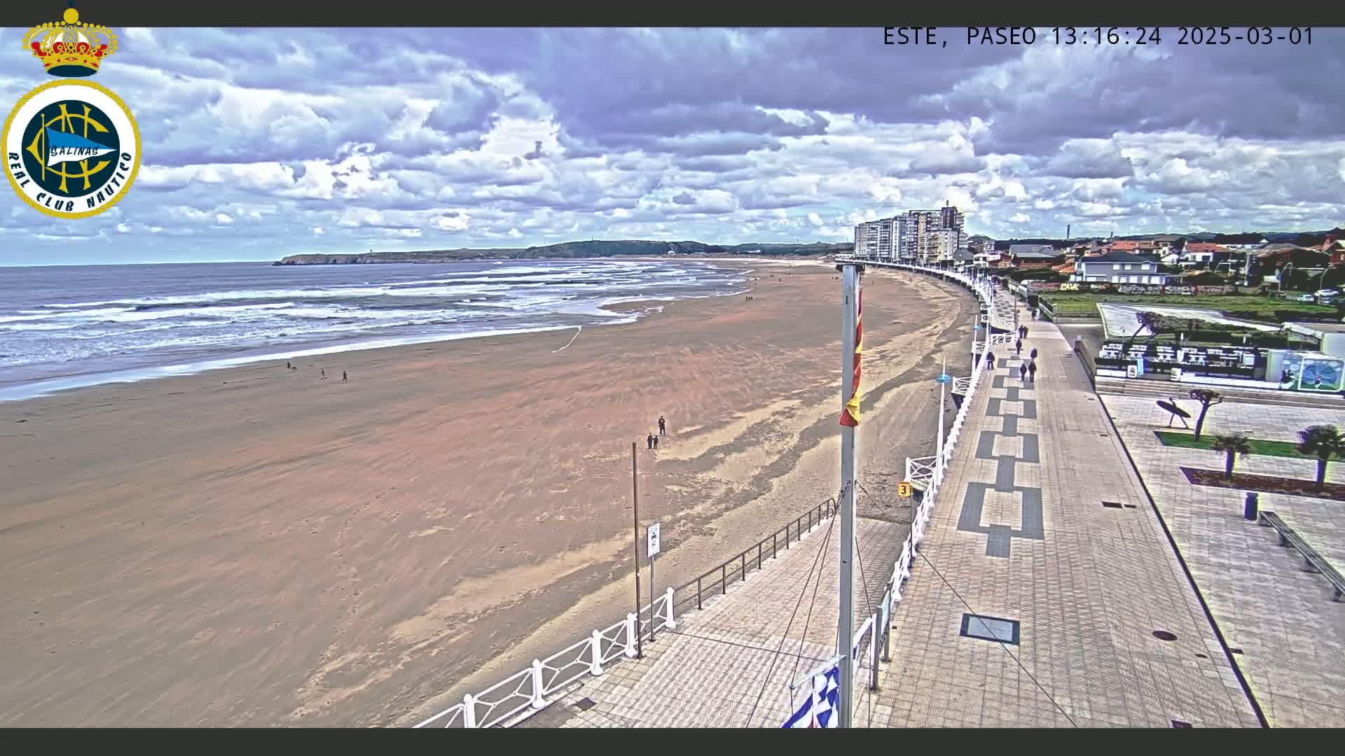 A sandy beach with moderate waves under a partly cloudy sky is bordered by a paved promenade with people walking along it, and buildings are visible in the background.