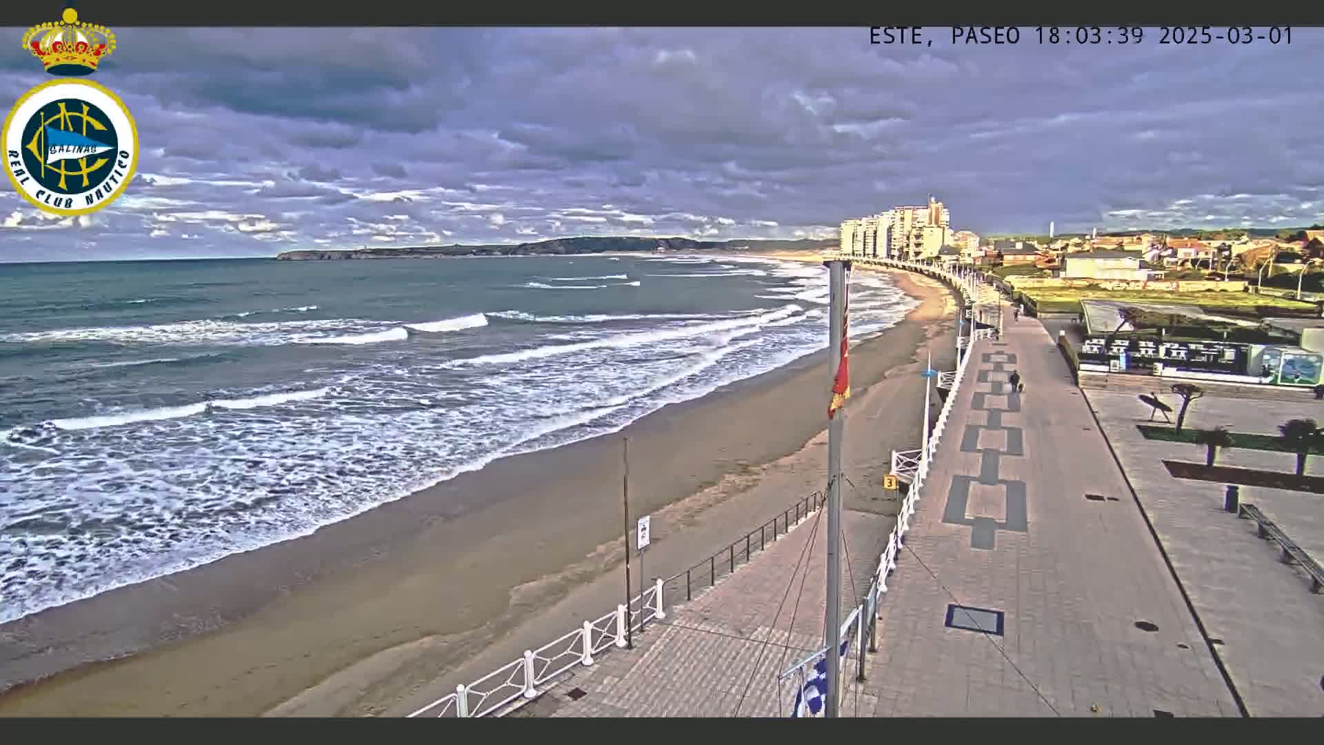 An overcast day shows a beach with moderate waves lapping a sandy shore, a paved walkway leading to buildings, and a few people walking along the path.