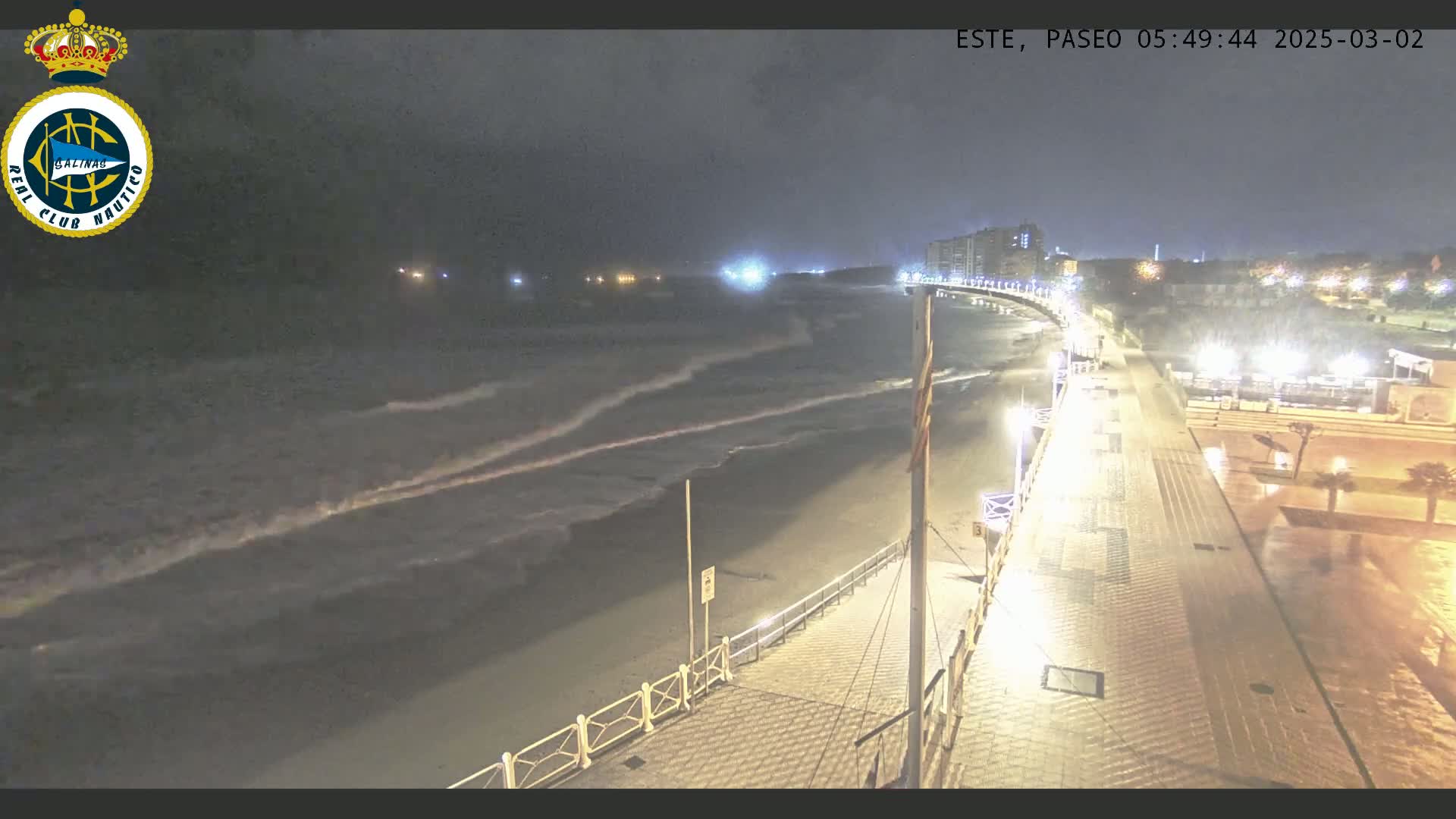 A nighttime view of a rain-slicked boardwalk overlooking a beach with moderate waves under a dark sky.