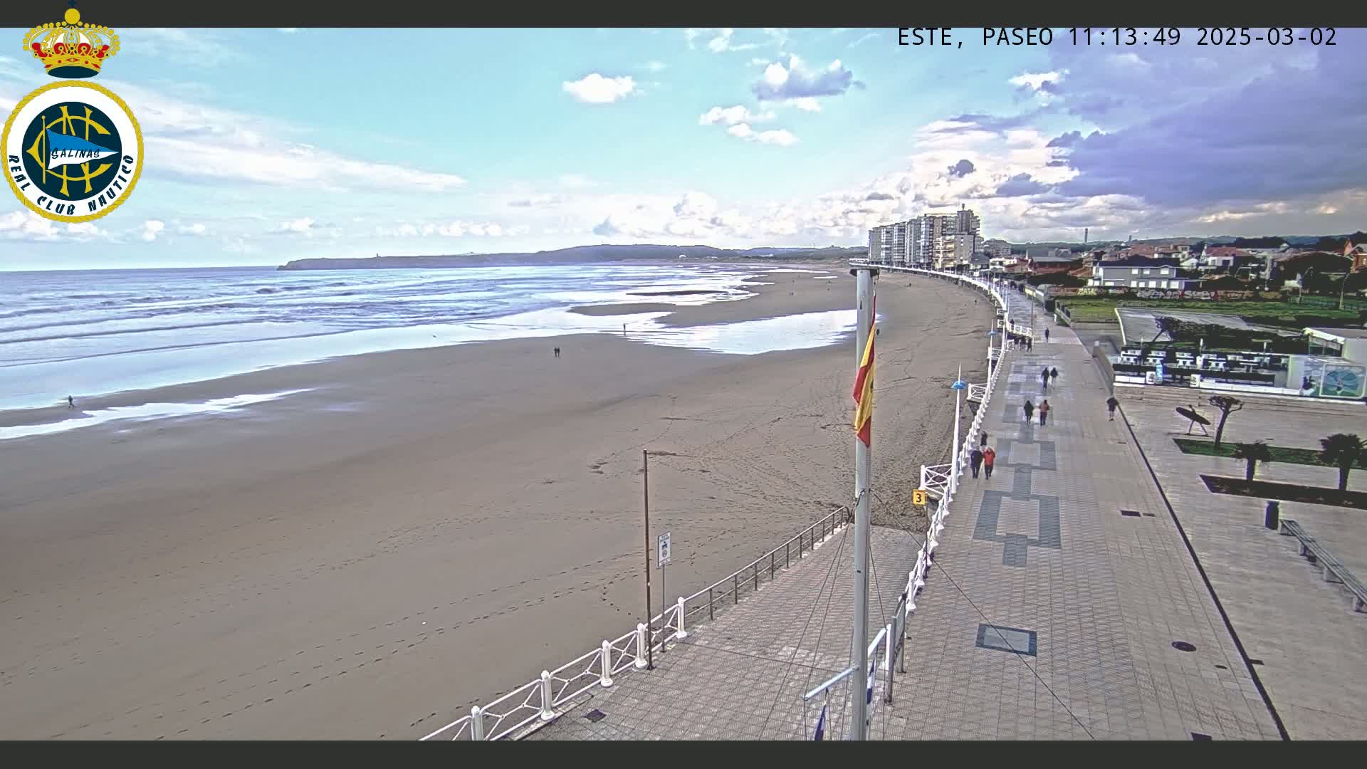 A sandy beach with gentle waves under a partly cloudy sky is bordered by a paved walkway with people strolling, and buildings are visible in the distance.