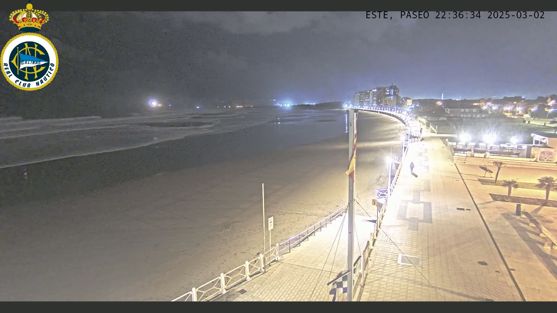 A nighttime view shows a mostly empty sandy beach with gentle waves under a dark sky, a boardwalk with lights lining a paved pathway next to the beach and buildings in the distance.