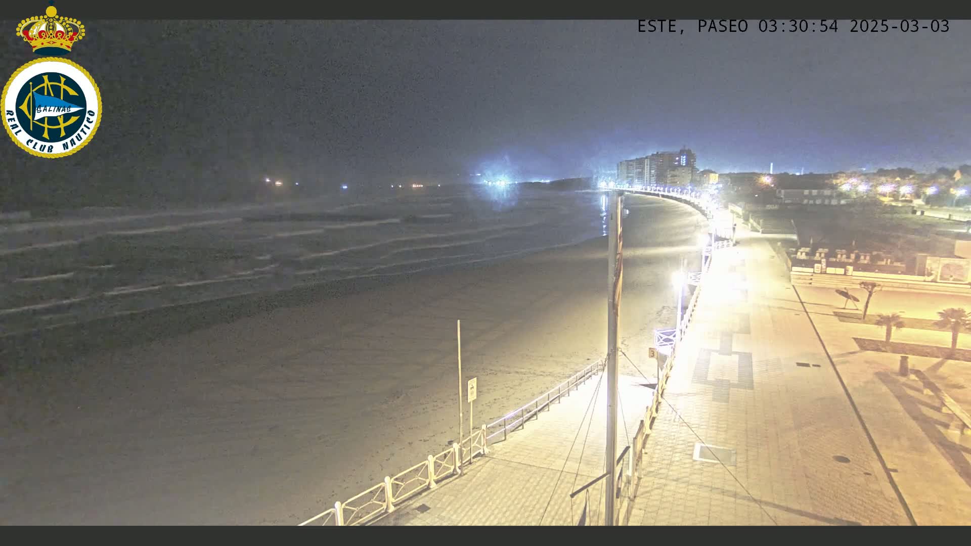 A nighttime view of a beach with gentle waves, a paved walkway lined with lights, and buildings in the distance under a dark sky.