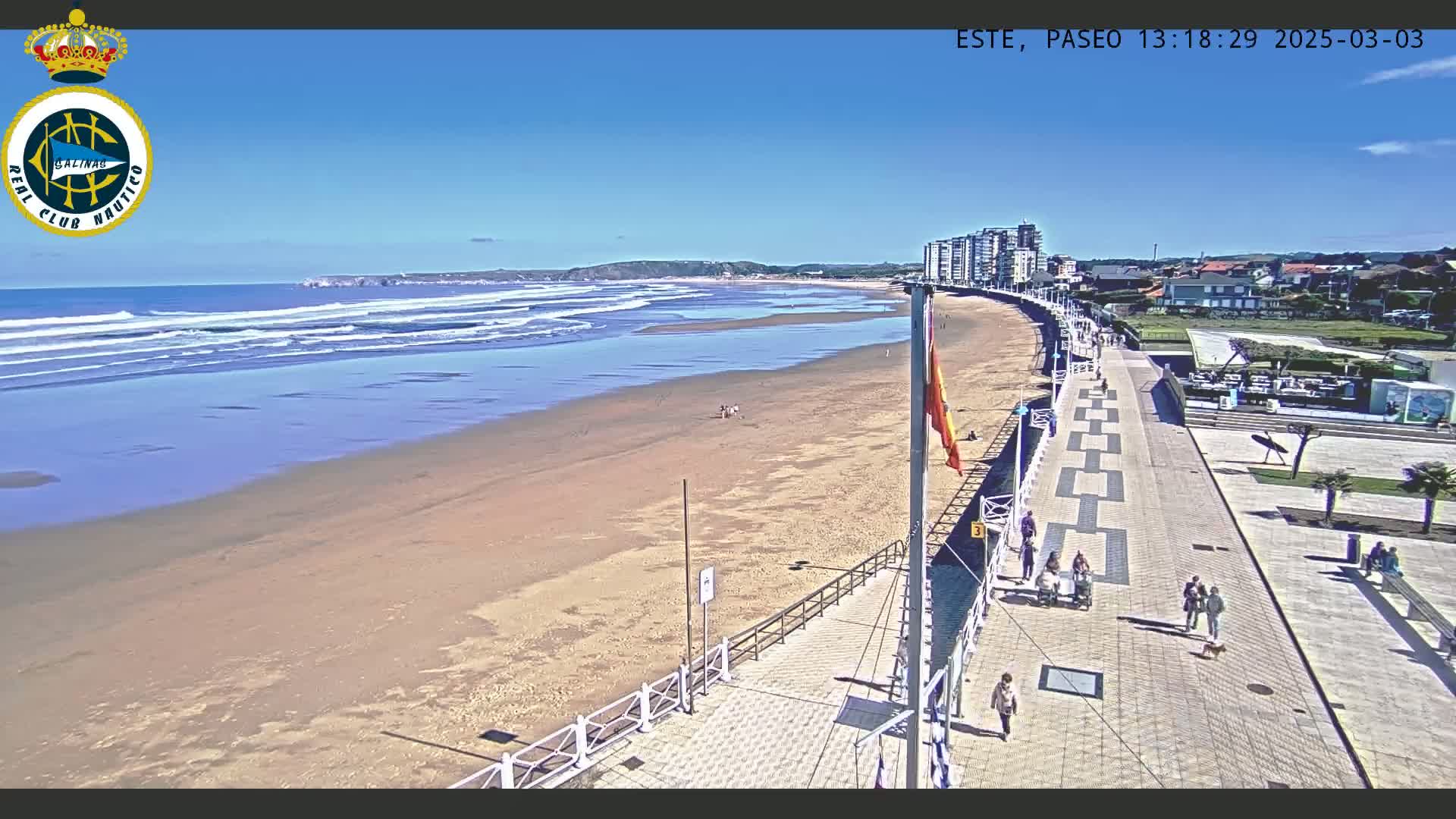 A sandy beach with gentle waves under a clear blue sky is bordered by a paved boardwalk with people walking, and buildings are visible in the background.