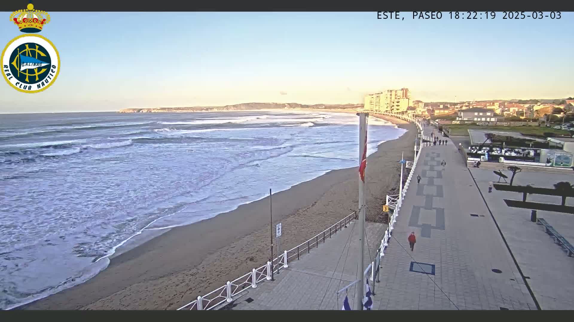 A sandy beach with gentle waves under a clear, sunny sky is bordered by a paved walkway with a few pedestrians, leading to buildings in the distance.