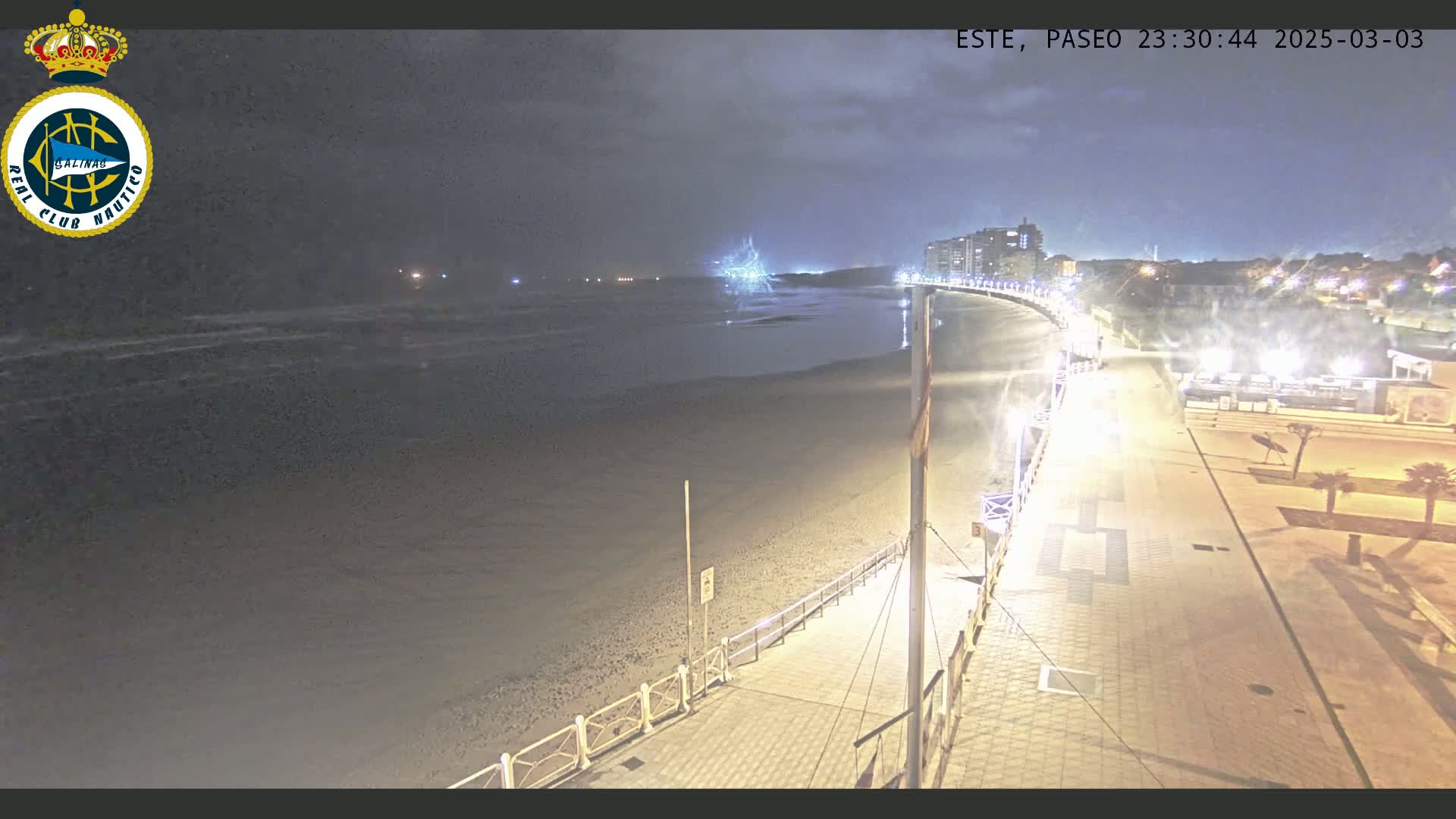 A nighttime view of a beach and boardwalk, with the ocean appearing calm under a partly cloudy sky.