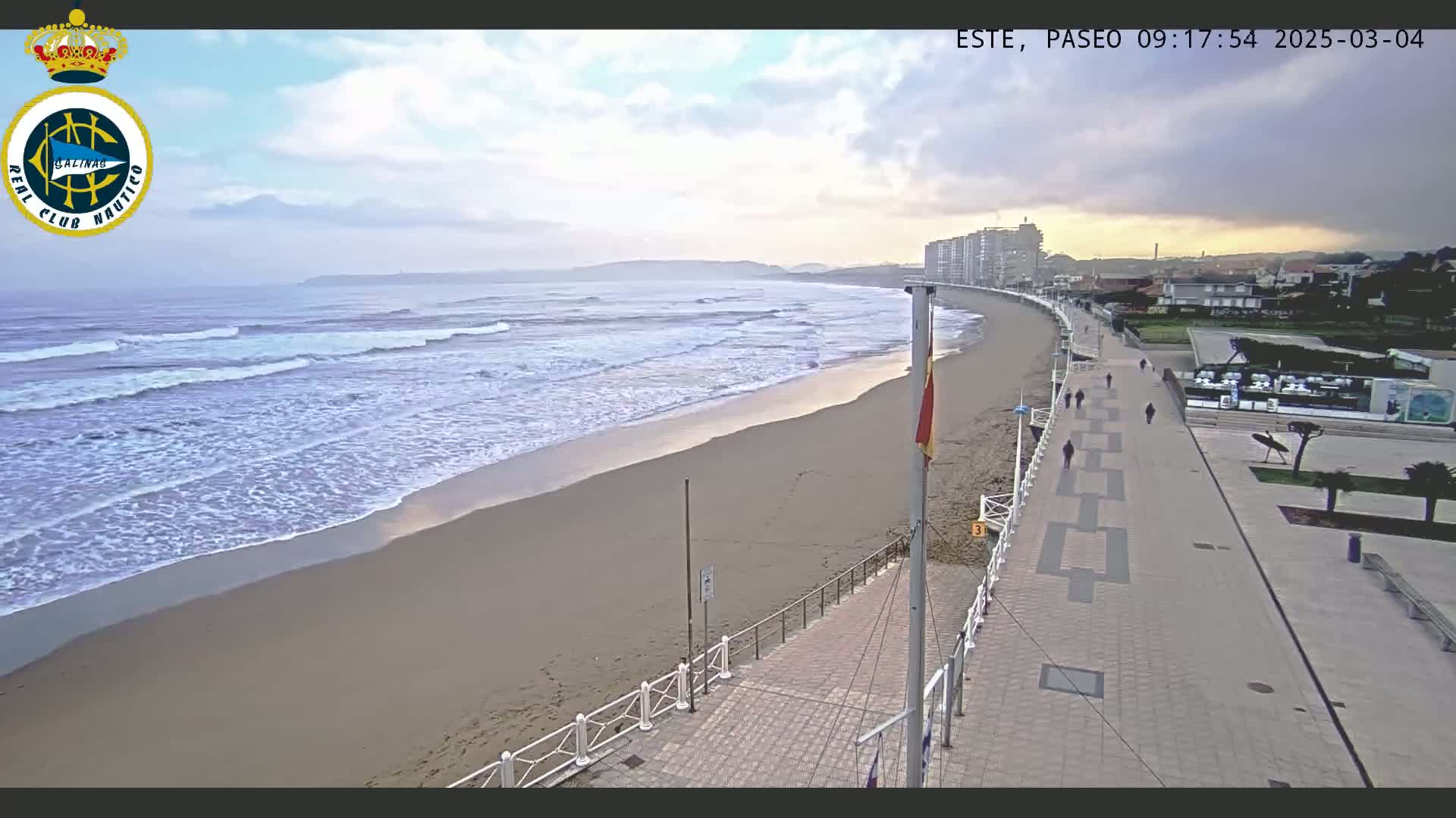 A sandy beach with gentle waves under a partly cloudy sky is next to a paved walkway with a few pedestrians, bordered by buildings and green spaces.