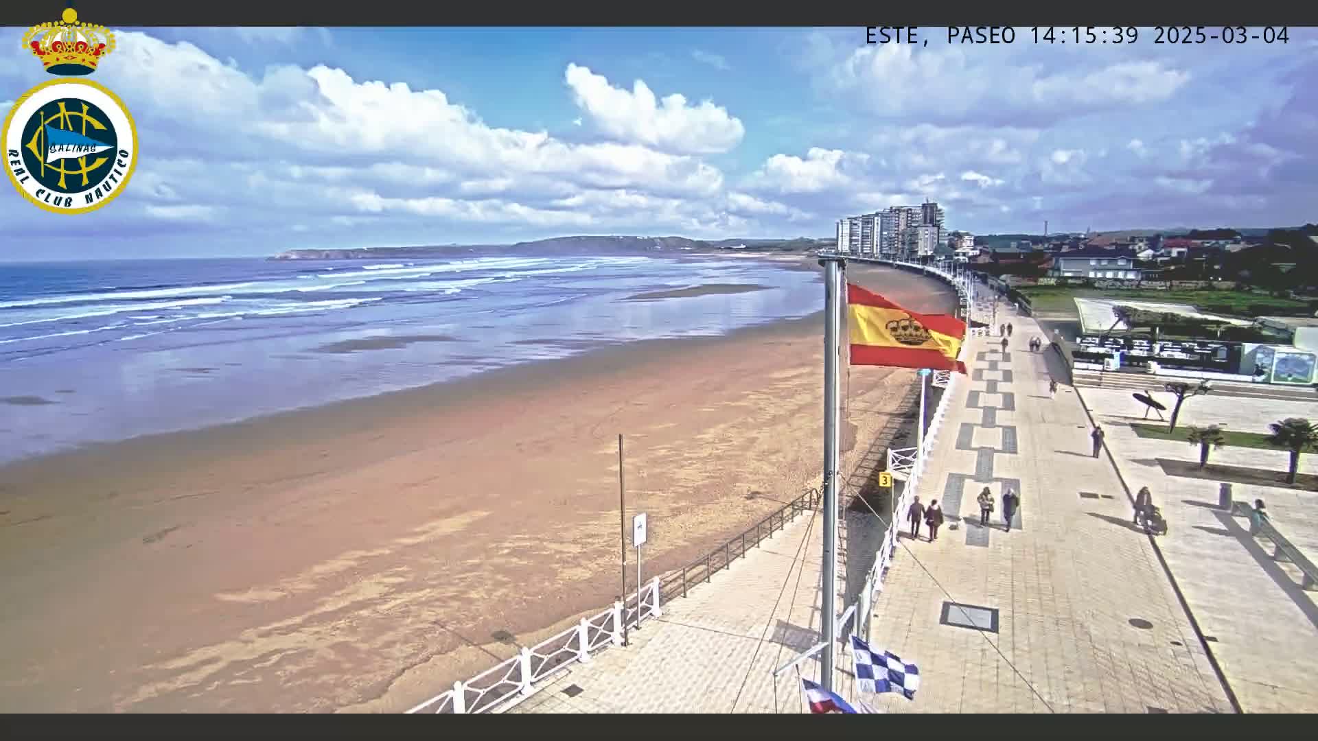 A wide sandy beach with small waves under a partly cloudy sky is bordered by a paved walkway with people strolling along it, and buildings are visible in the distance.