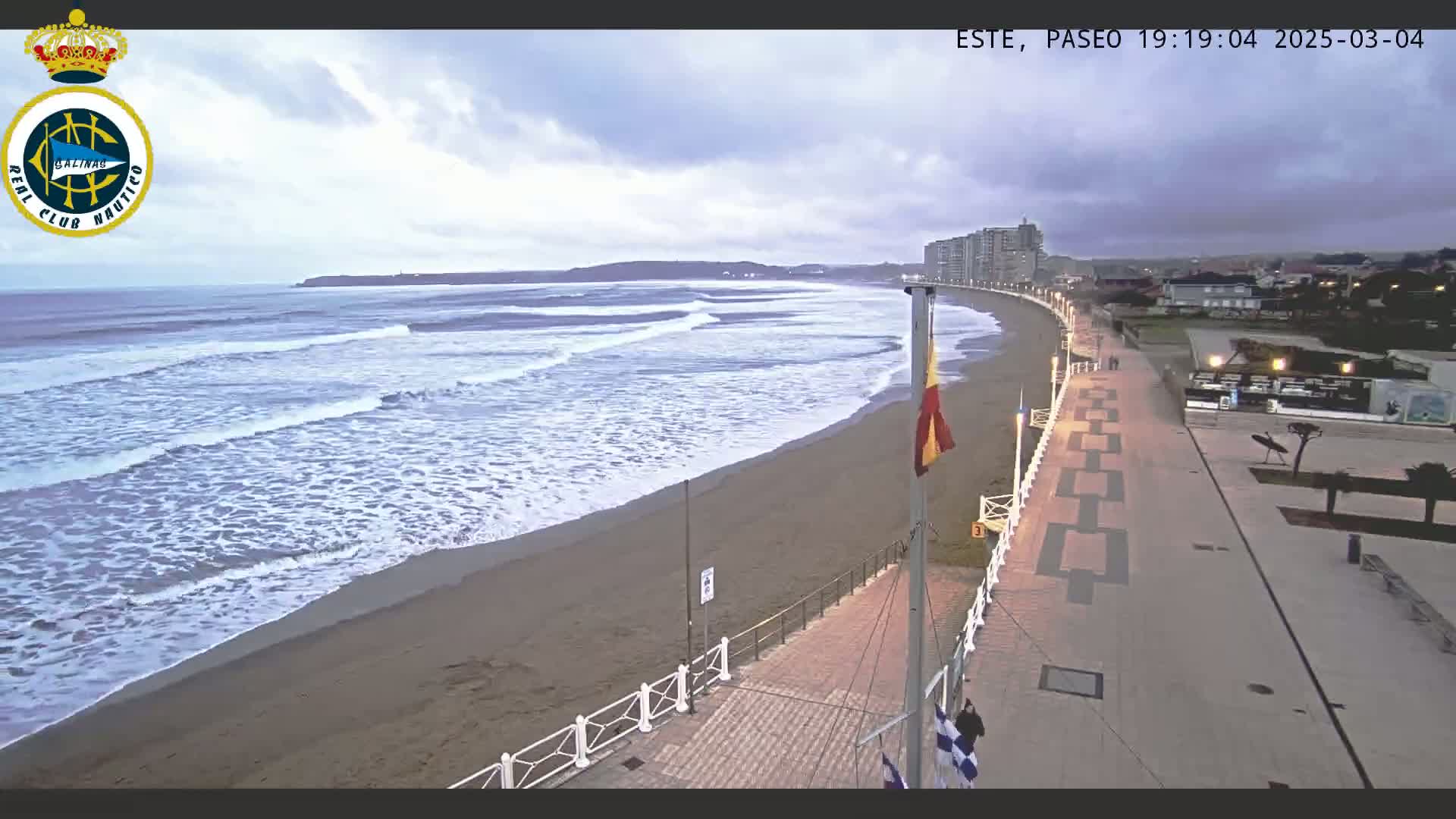 A sandy beach with moderate waves under an overcast sky is bordered by a paved walkway and buildings.