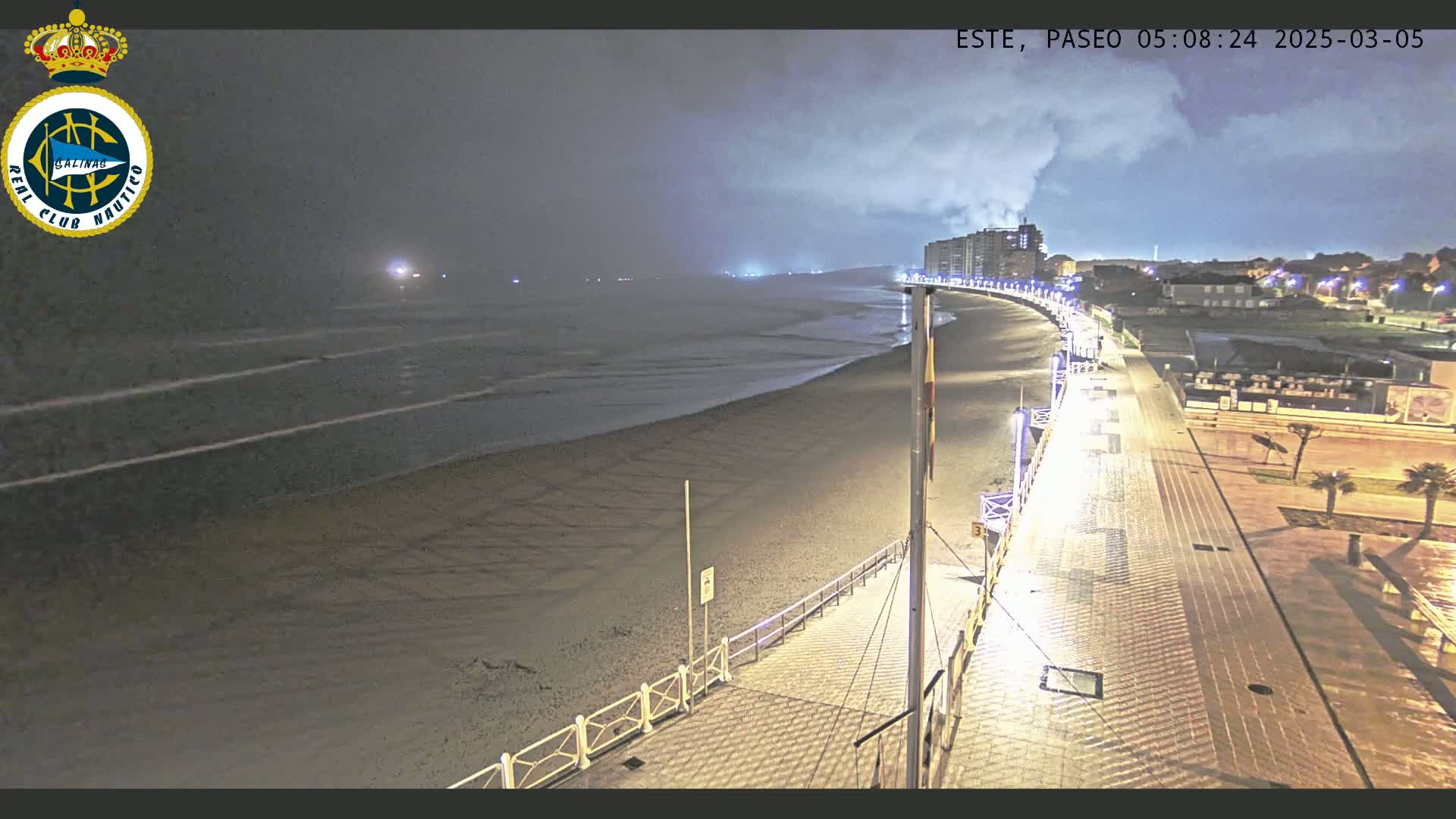 A nighttime view of a mostly empty beach and boardwalk, under a cloudy sky, with lights illuminating the paved walkway and buildings in the distance.