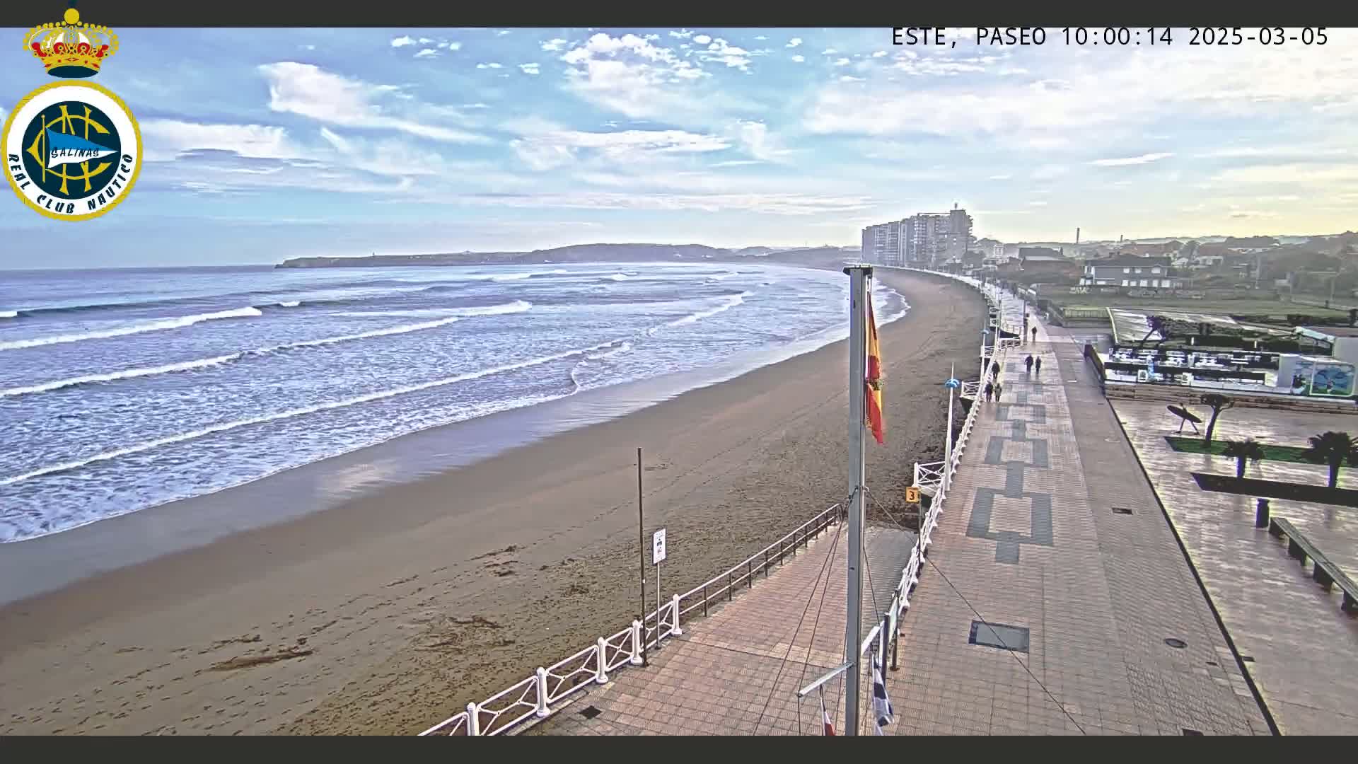 A sandy beach with gentle waves under a partly cloudy sky is bordered by a paved walkway lined with buildings and a few people walking.