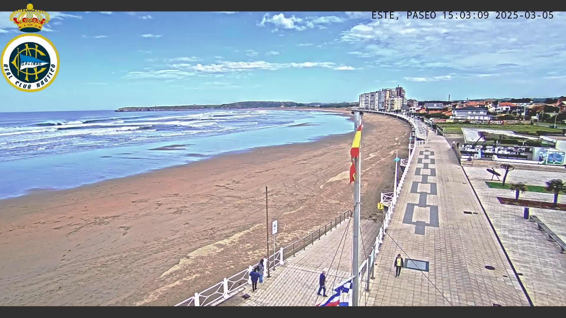 A sandy beach with small waves under a mostly sunny sky is bordered by a paved walkway leading to buildings, with a few people walking along the path.