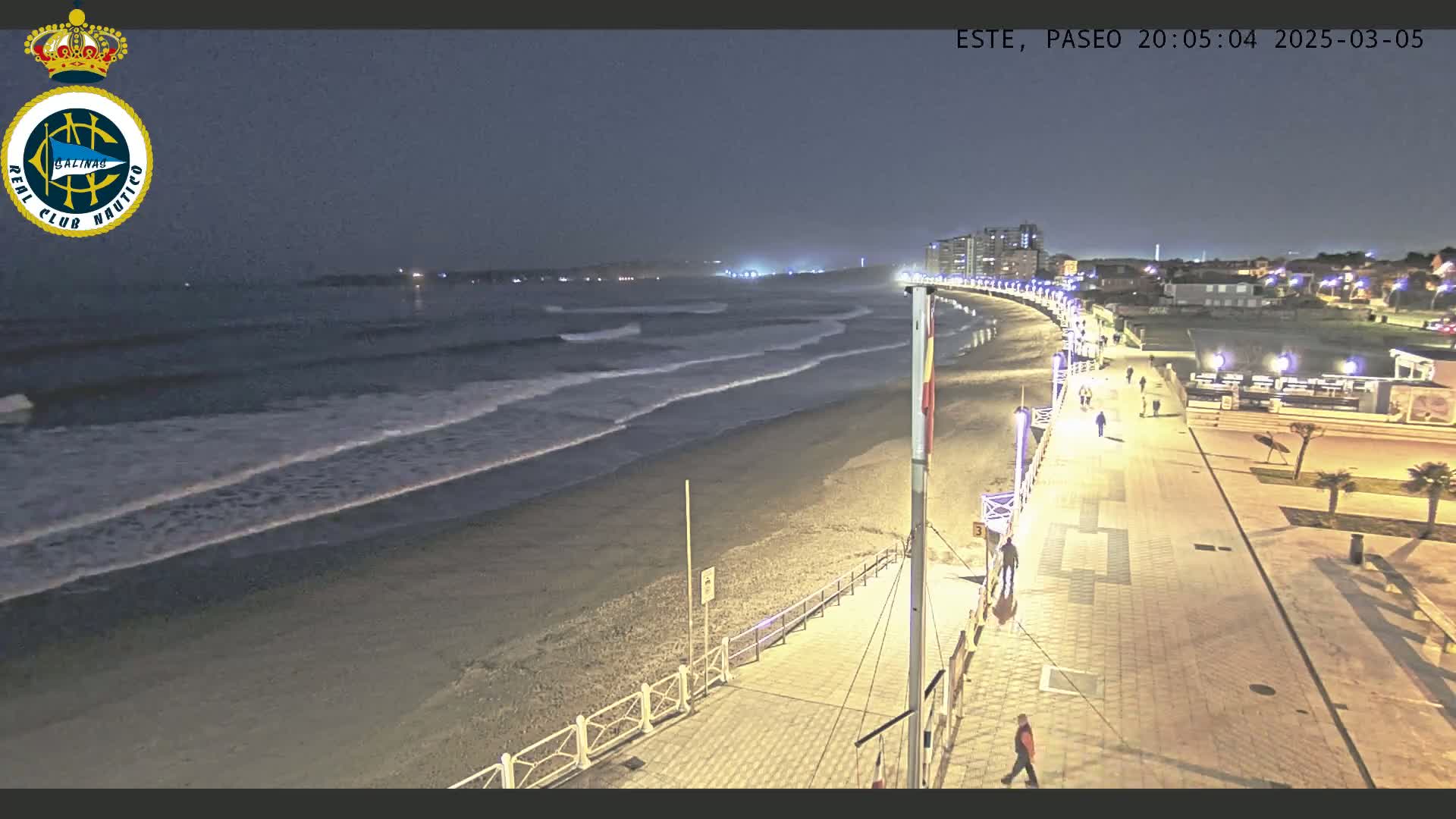 A nighttime beach scene shows gentle waves lapping the shore, a boardwalk with pedestrians, and buildings in the distance under a dark sky.
