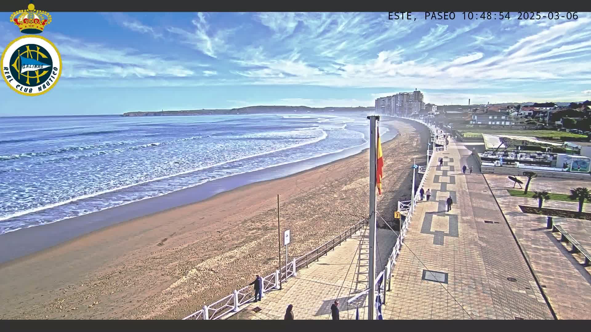 A sandy beach with gentle waves under a mostly sunny sky is bordered by a paved boardwalk with people walking, and buildings are visible in the distance.
