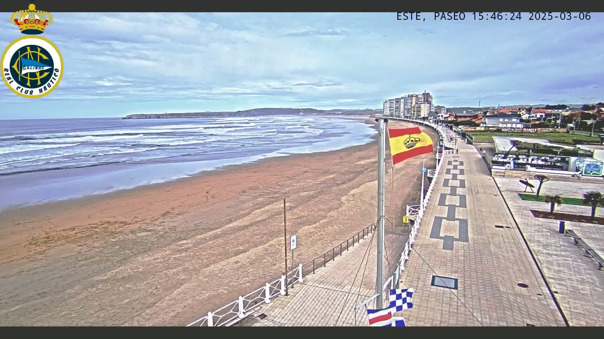 A sandy beach with moderate waves under a partly cloudy sky is viewed from a paved boardwalk, alongside buildings and a Spanish flag.