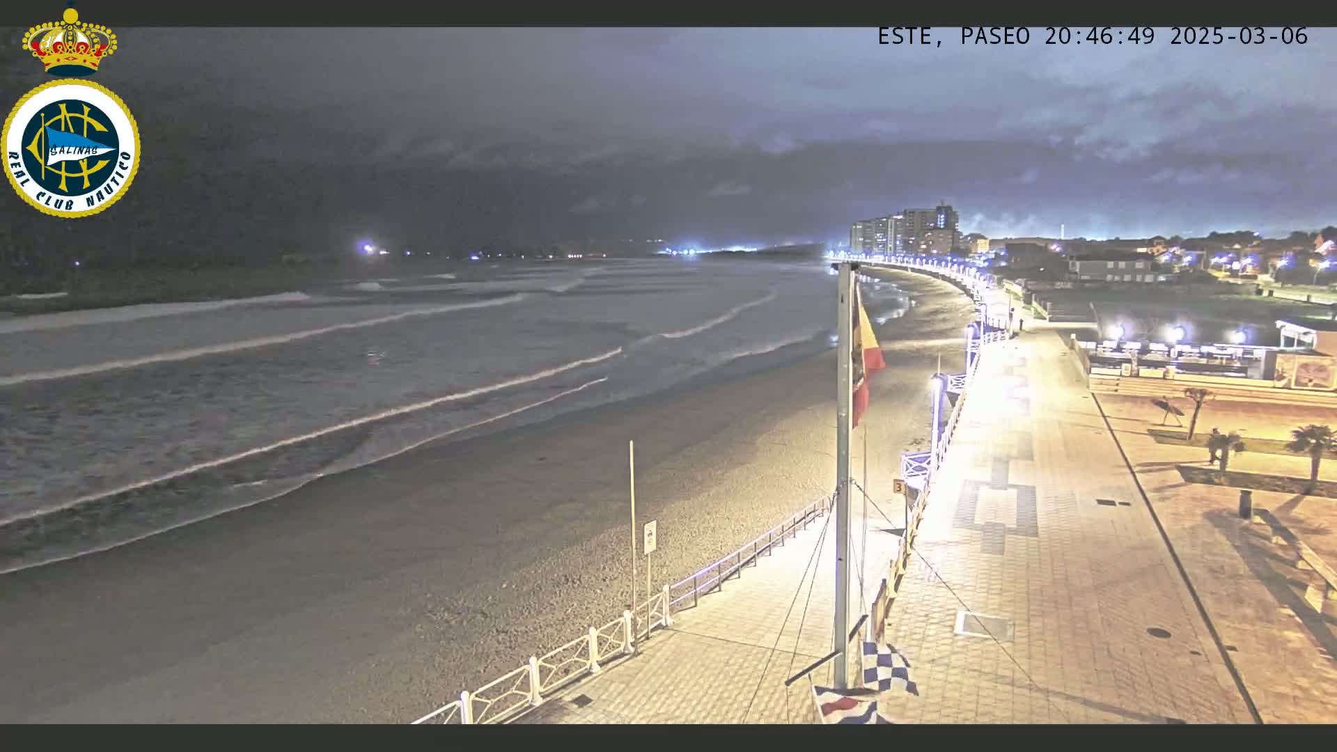 A nighttime view of a beach with gentle waves rolling onto the shore, a paved walkway lined with lights, and buildings visible in the distance under a cloudy sky.