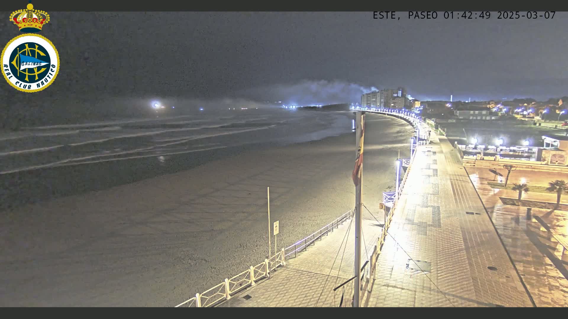 A nighttime view of a mostly empty beach with moderate waves under a cloudy sky, next to a paved boardwalk with lights and buildings in the distance.