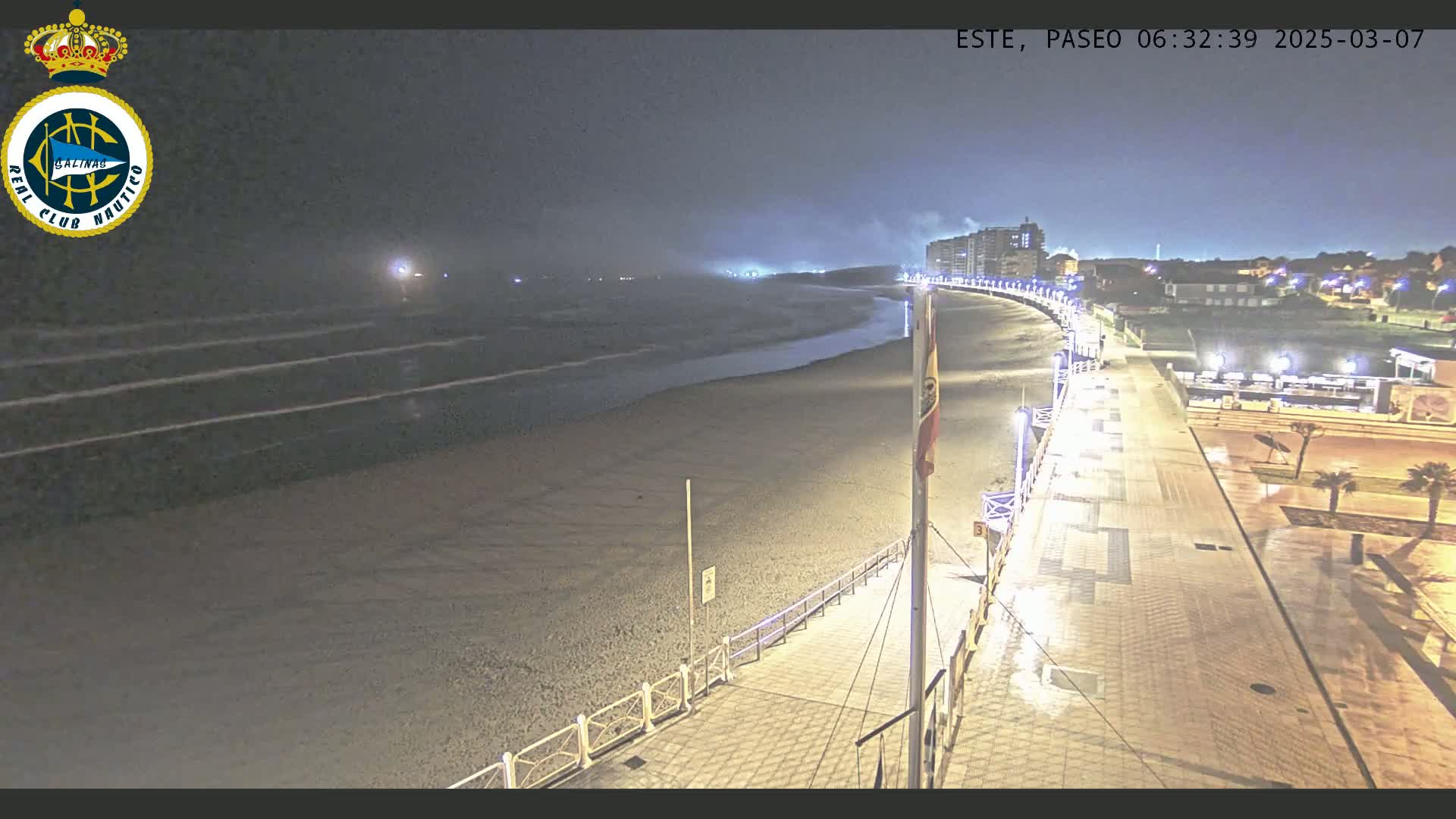 A nighttime view of a mostly empty beach and boardwalk, with calm waves and hazy weather conditions.