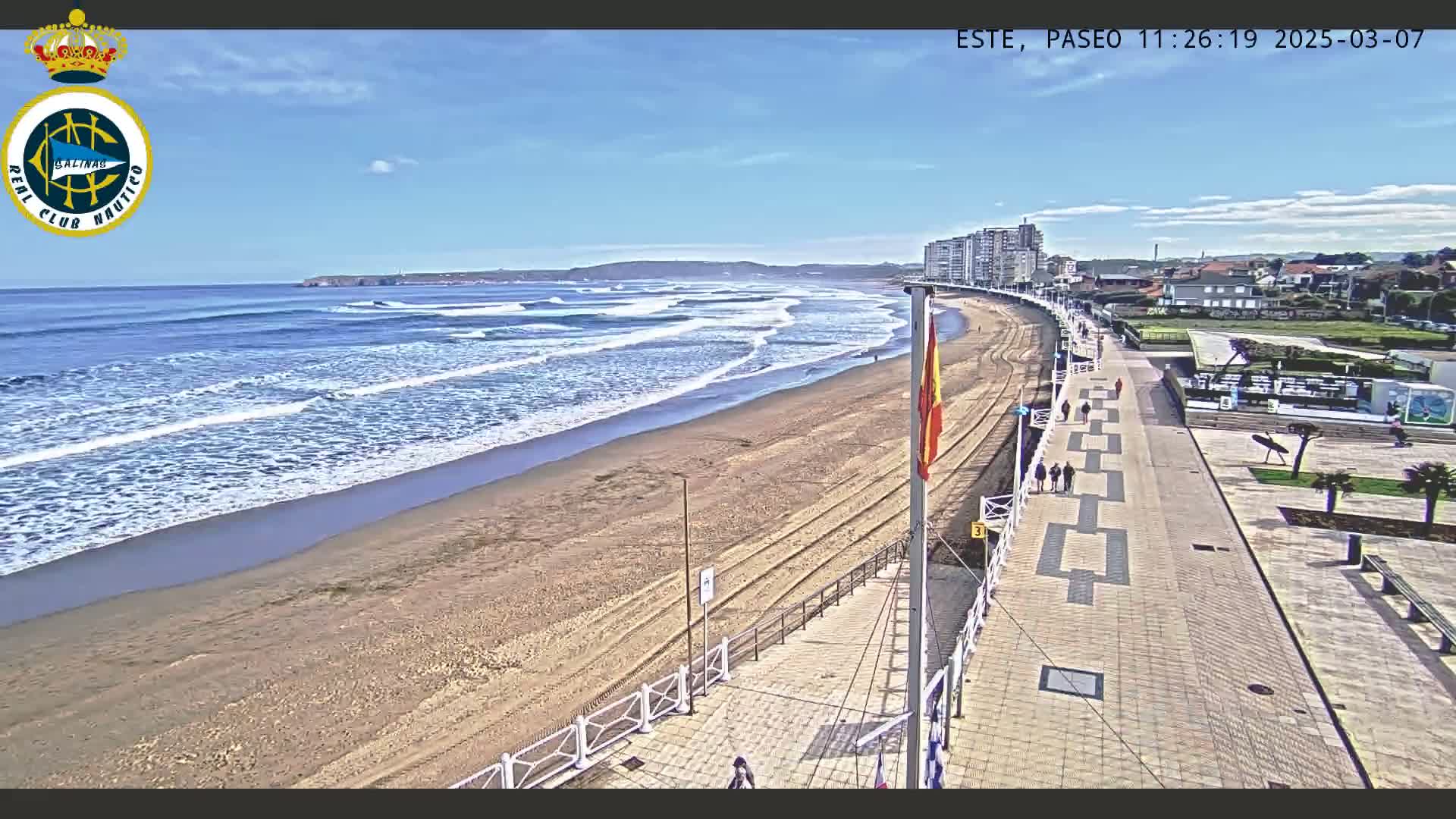 A sandy beach with gentle waves under a clear blue sky is bordered by a paved boardwalk and buildings, with several people walking along the promenade.