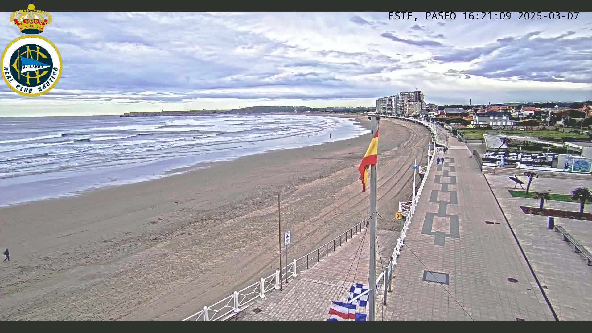 A sandy beach with small waves under a partly cloudy sky is adjacent to a paved boardwalk lined with buildings.