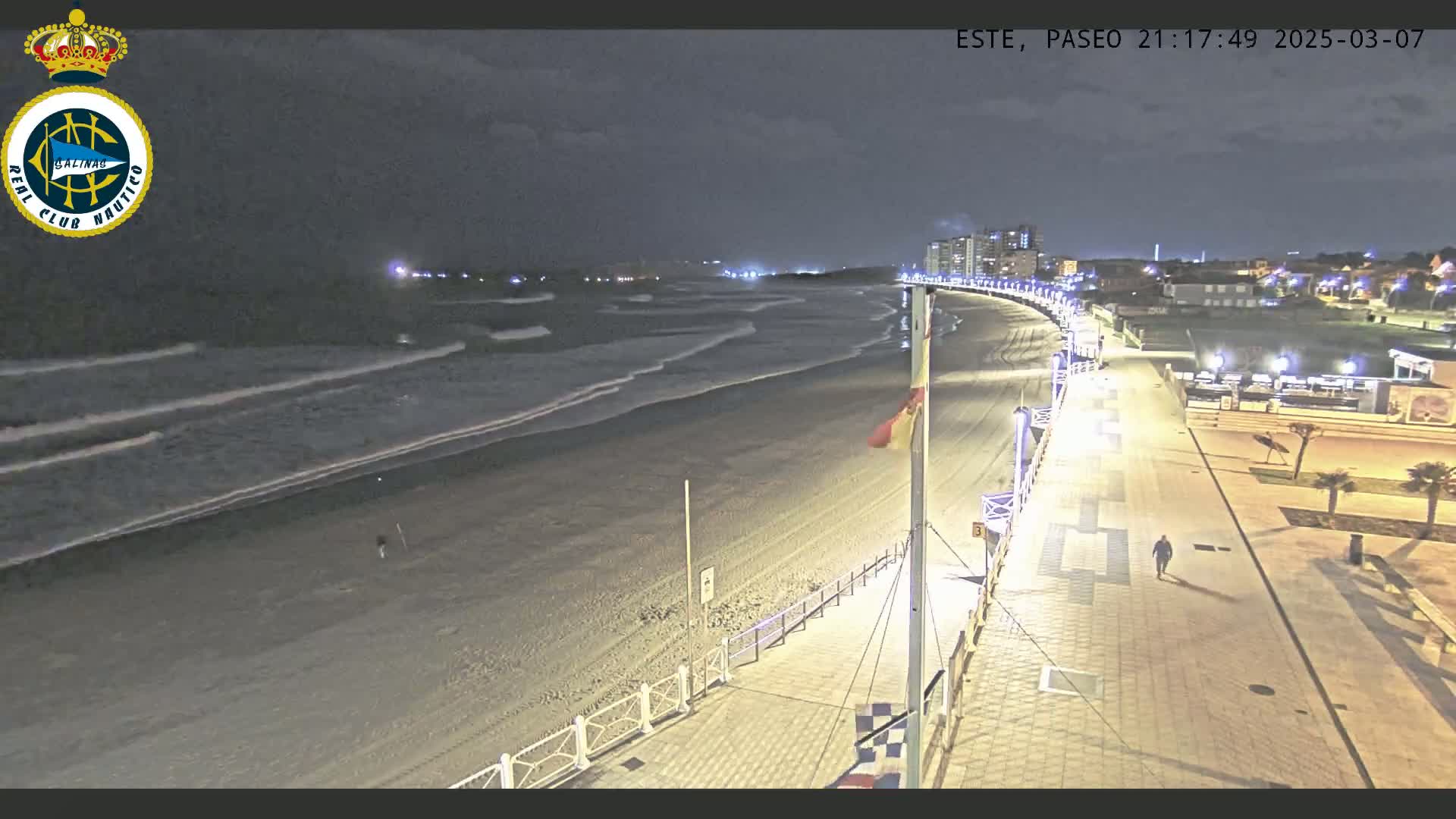A nighttime view shows a mostly empty beach with gentle waves, a paved boardwalk with lights, and a few buildings in the distance under a dark, cloudy sky.