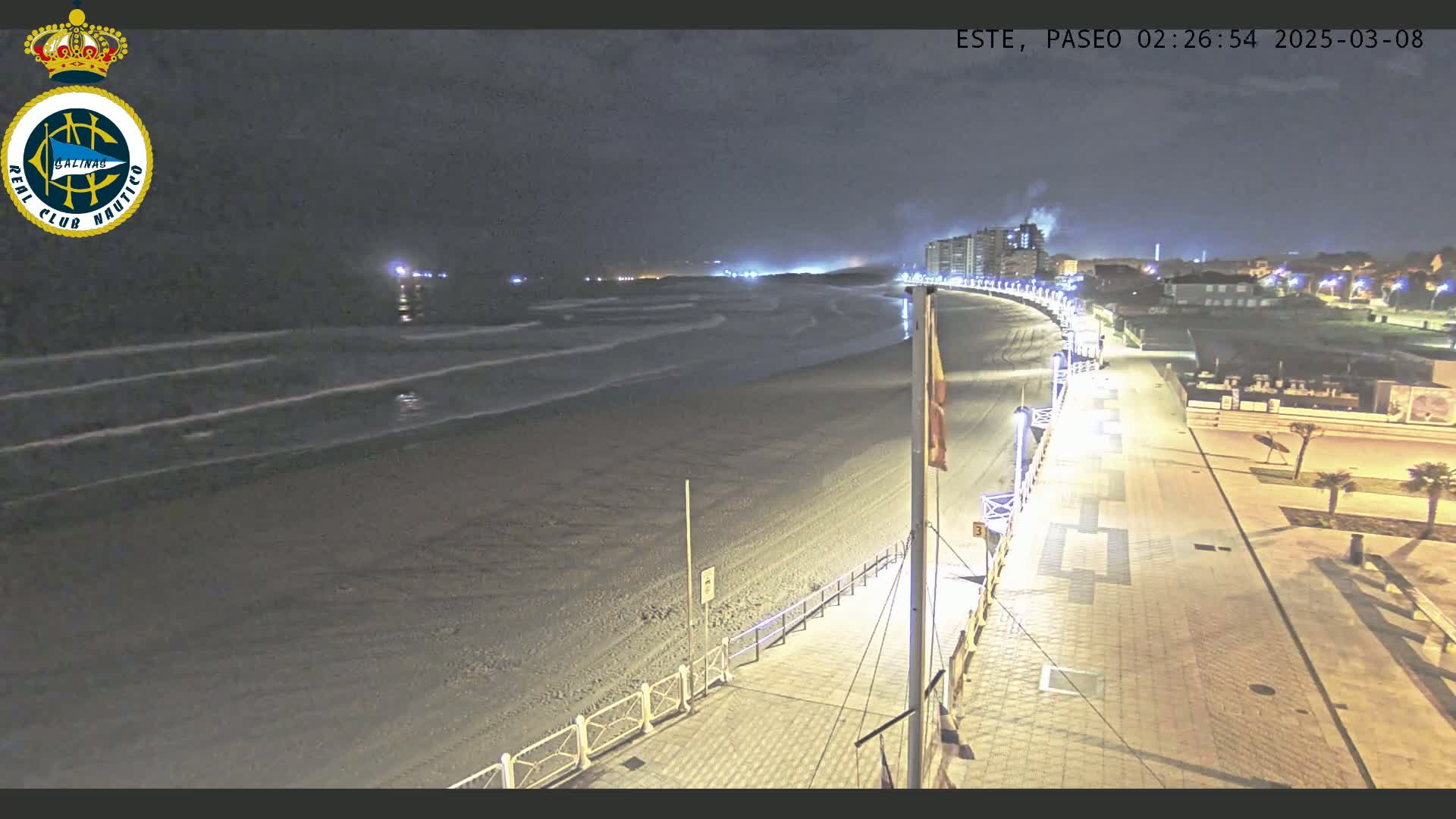 A nighttime view of a mostly empty beach with gentle waves, a paved walkway lined with lights, and buildings in the distance under a dark sky.