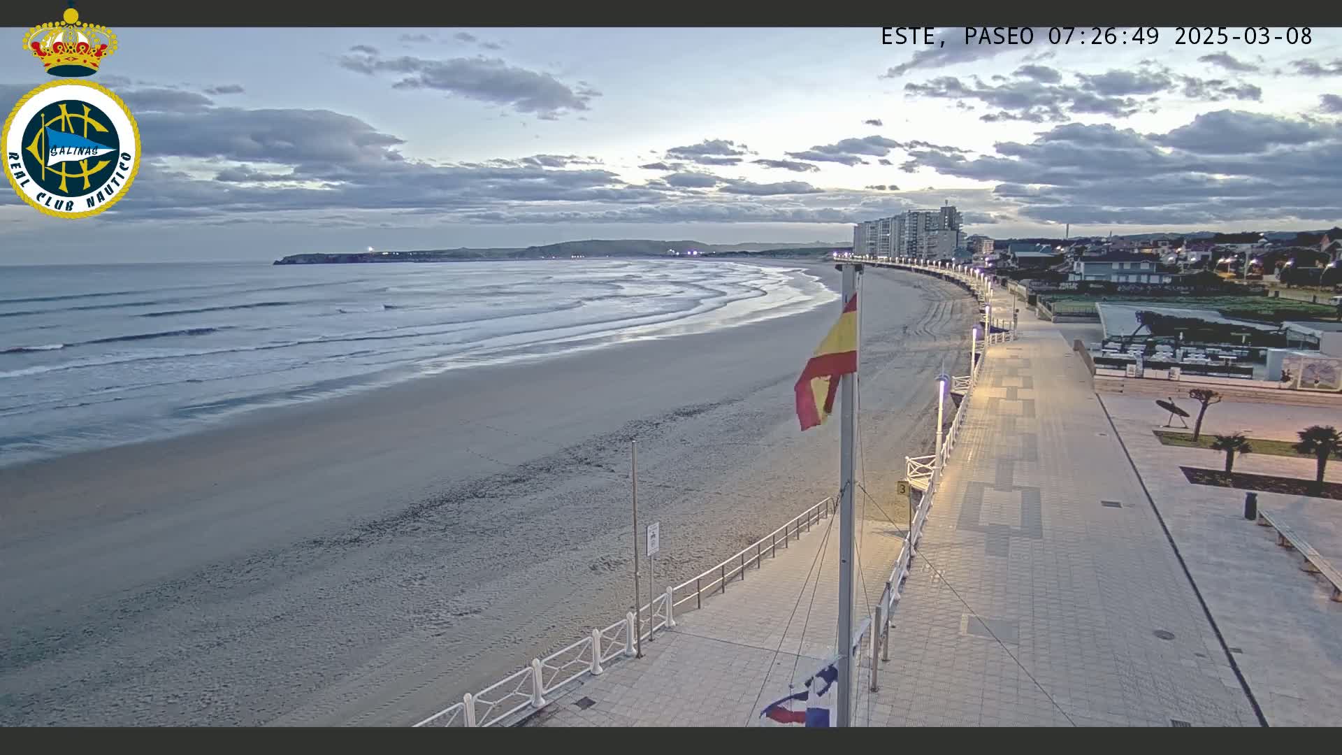 An overcast sunrise illuminates a sandy beach with gentle waves, a boardwalk, and buildings in the distance.