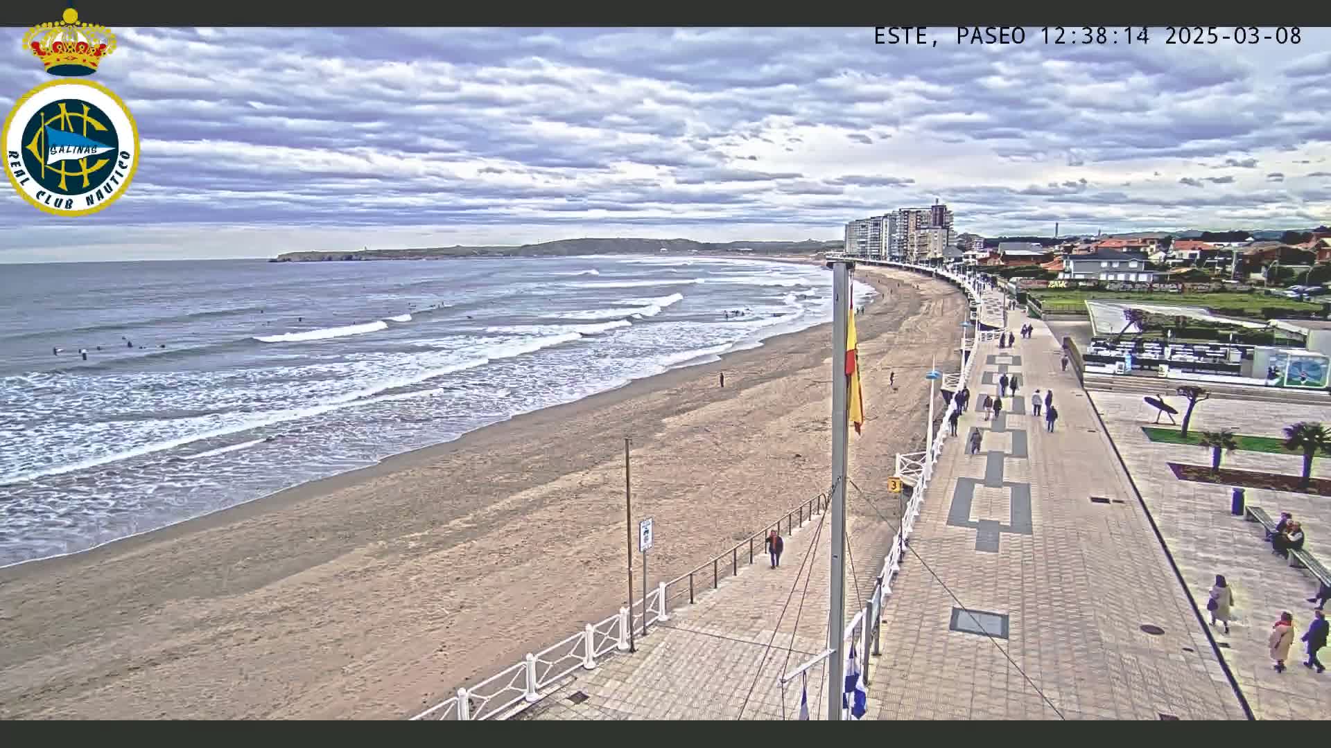 A sandy beach with moderate waves under a partly cloudy sky is viewed from above a paved boardwalk with people walking along it and buildings in the background.
