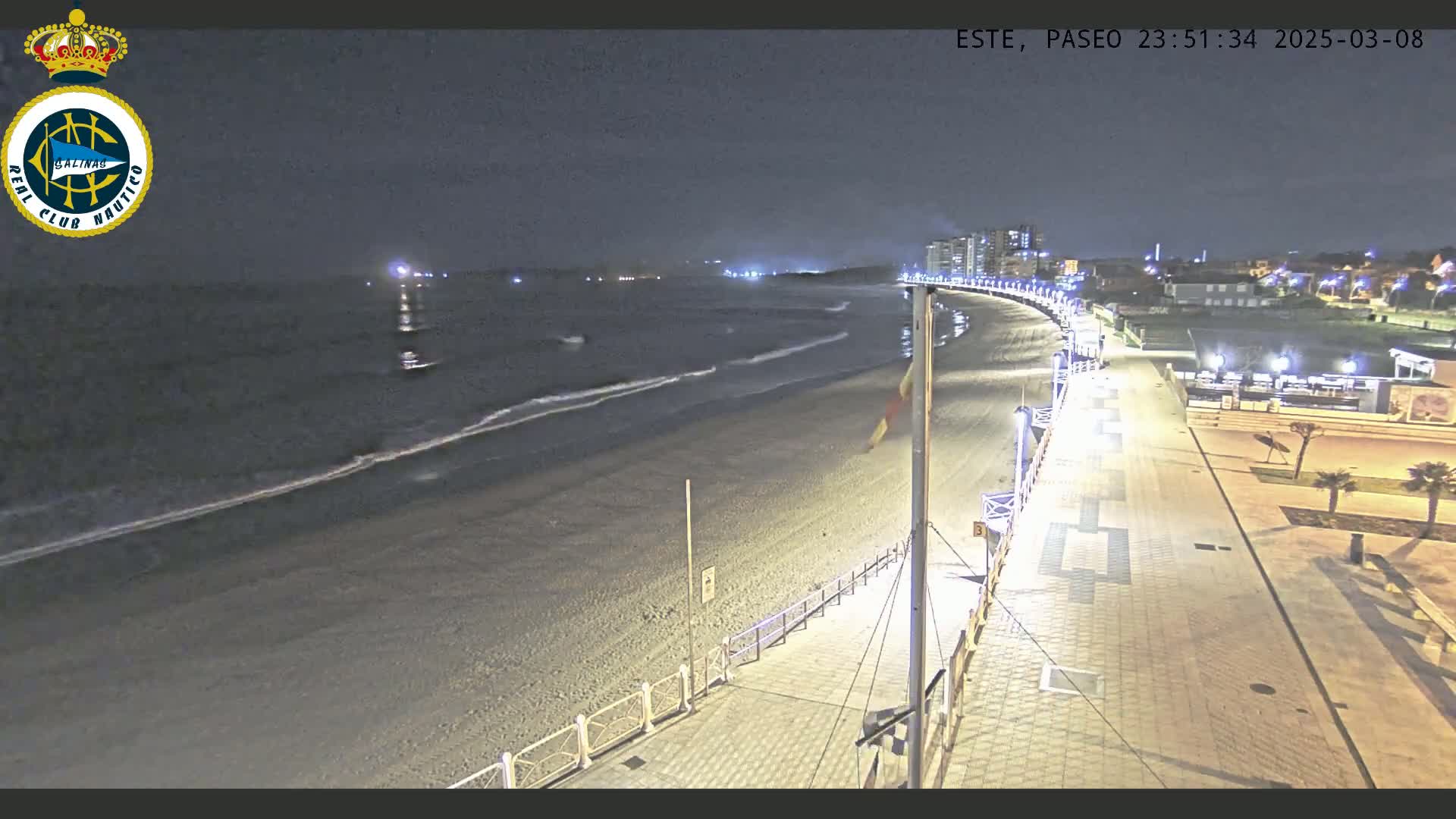 A nighttime view of a beach with gentle waves lapping the shore, a paved walkway lined with lights, and buildings visible in the distance.