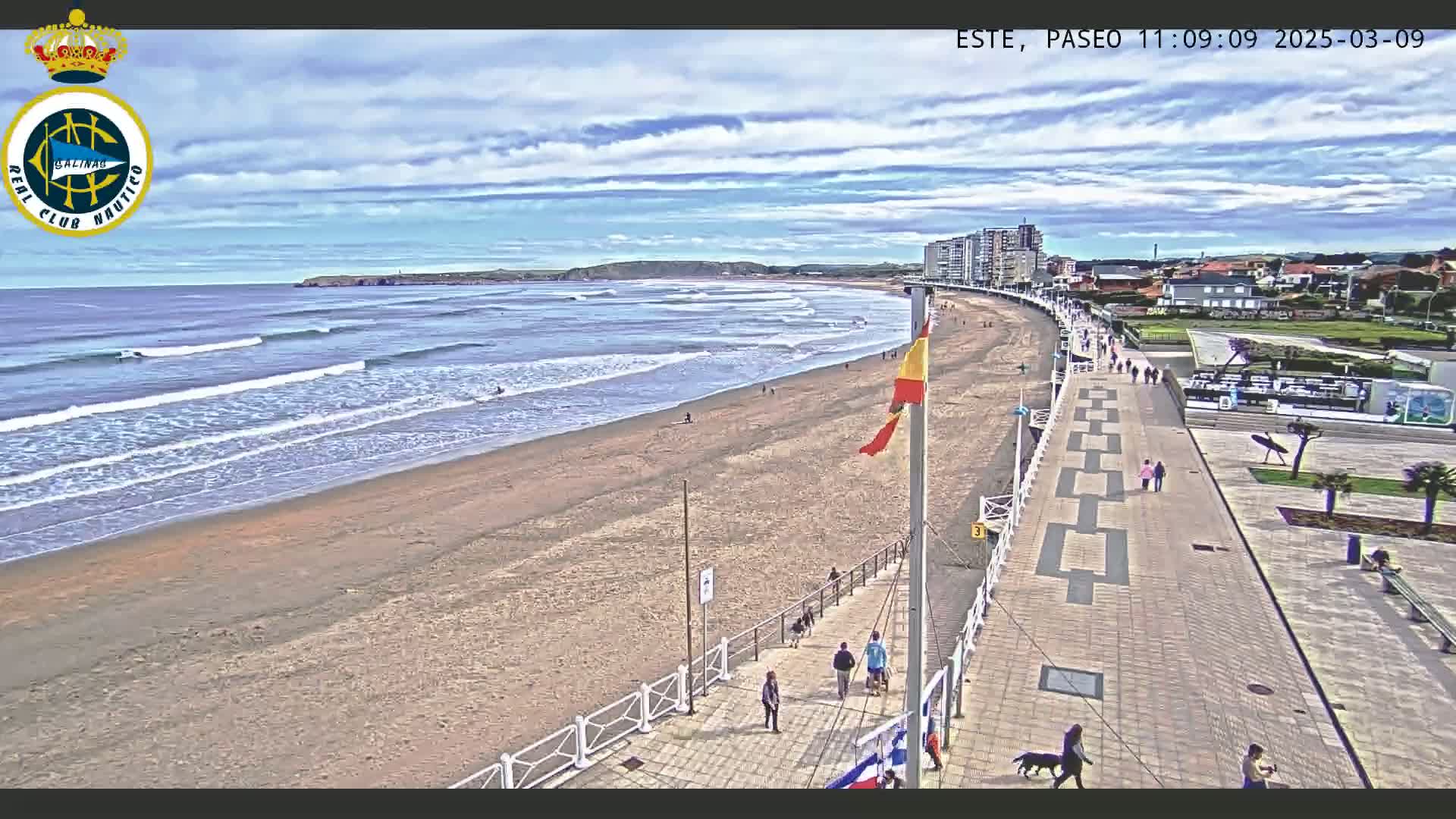 A sandy beach with gentle waves under a partly cloudy sky is bordered by a paved walkway with several pedestrians and dogs.