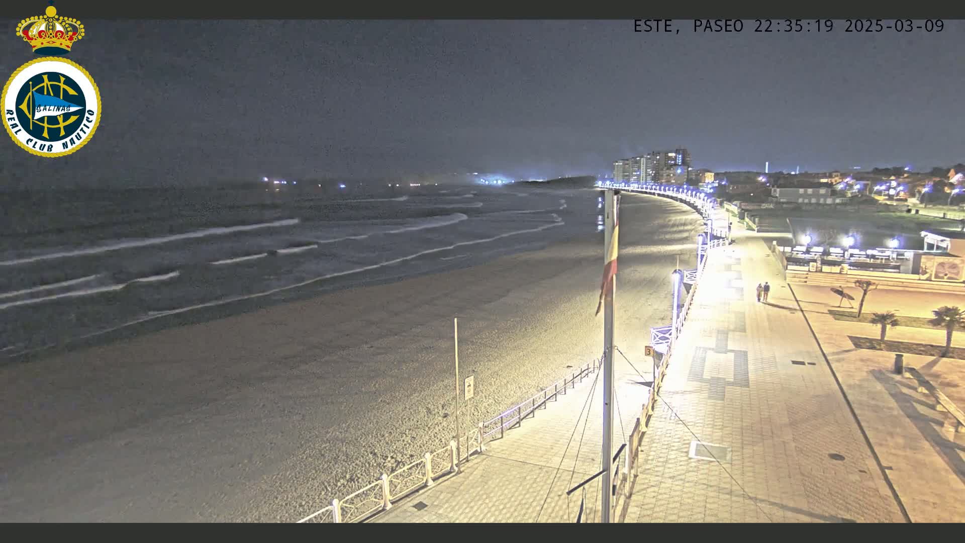 A nighttime, overhead view shows a sandy beach with gentle waves lapping the shore, adjacent to a paved boardwalk with lights illuminating the path and a few people walking.