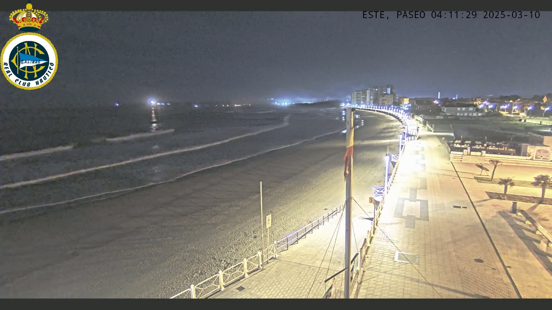 A nighttime view of a dark, sandy beach with gentle waves lapping the shore, a paved walkway lined with lights, and buildings visible in the distance.
