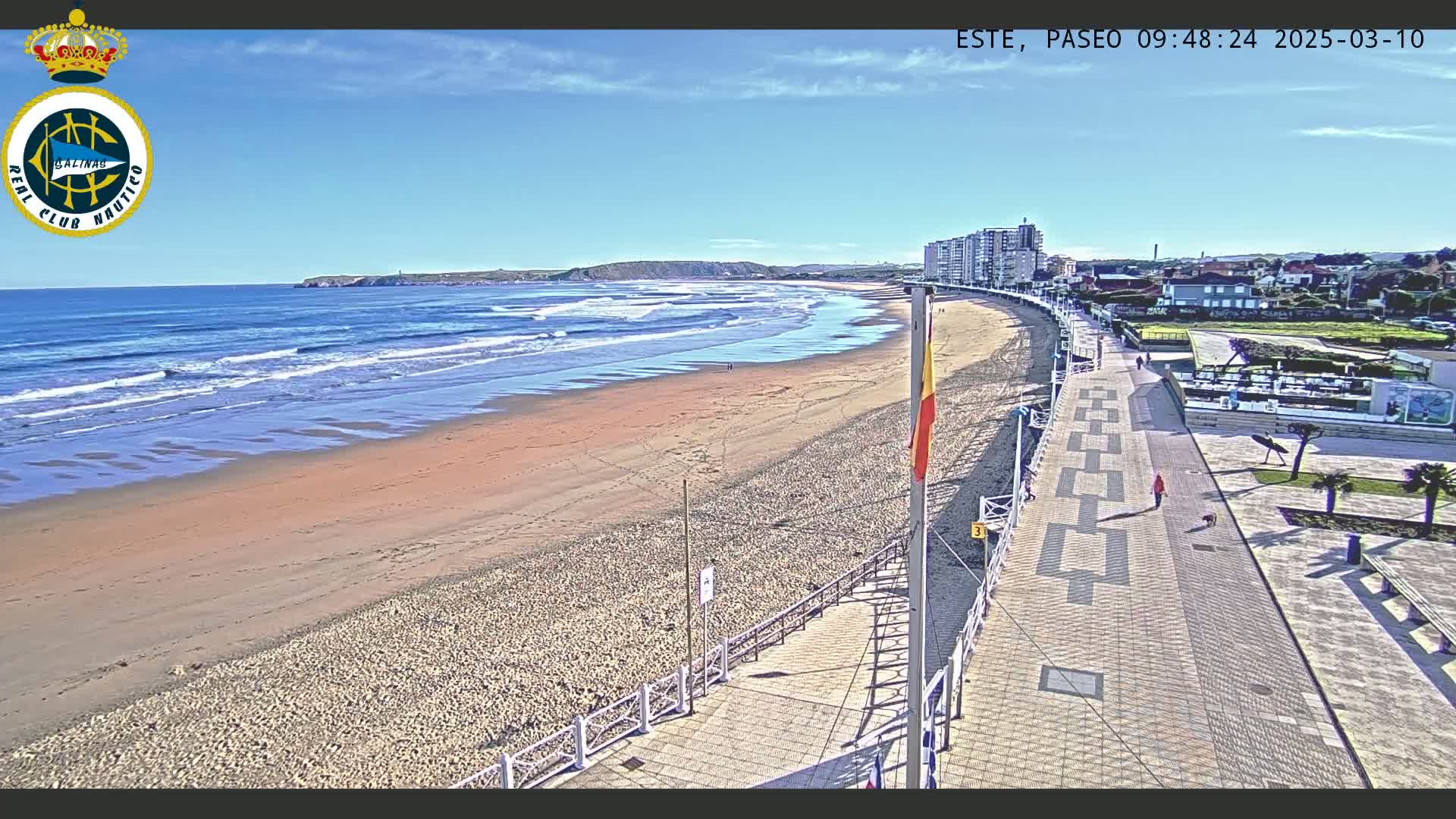 A sandy beach with gentle waves under a clear blue sky is bordered by a paved walkway leading to buildings and a small town.