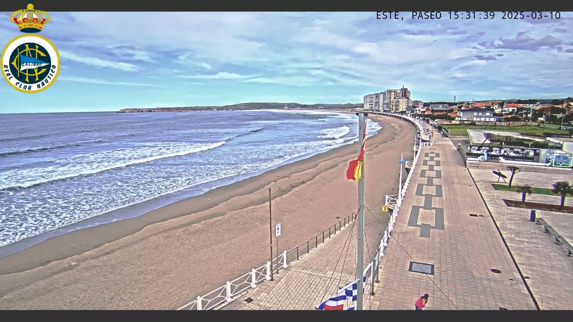 A sandy beach with gentle waves under a partly cloudy sky is bordered by a paved walkway leading to buildings along the coast.
