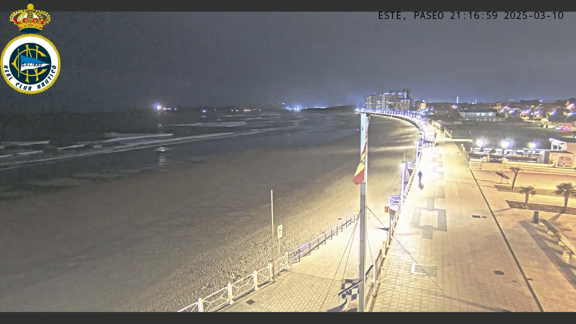 A nighttime view of a beach with gentle waves, a paved walkway lined with lights, and buildings in the distance under a dark sky.