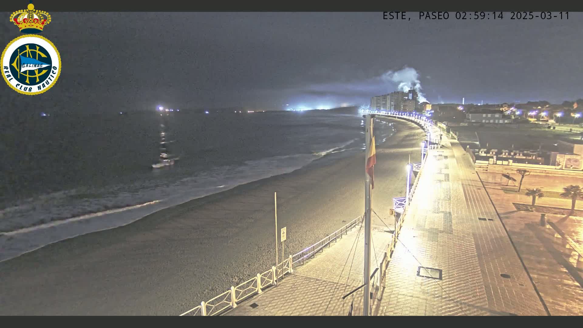 A nighttime view of a beach with gentle waves, a paved boardwalk lined with lights, and buildings in the background under a dark, cloudy sky.