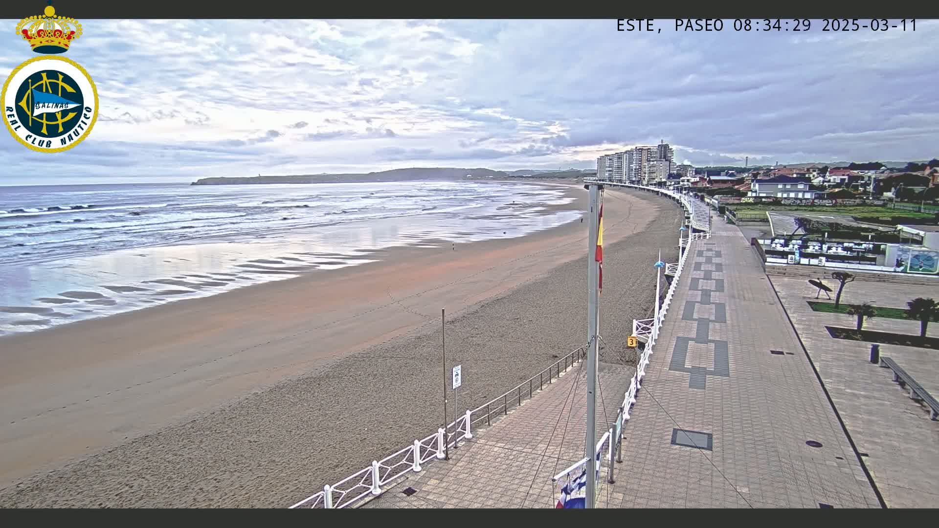 A sandy beach with gentle waves under a cloudy sky is bordered by a paved walkway leading to buildings and a town.