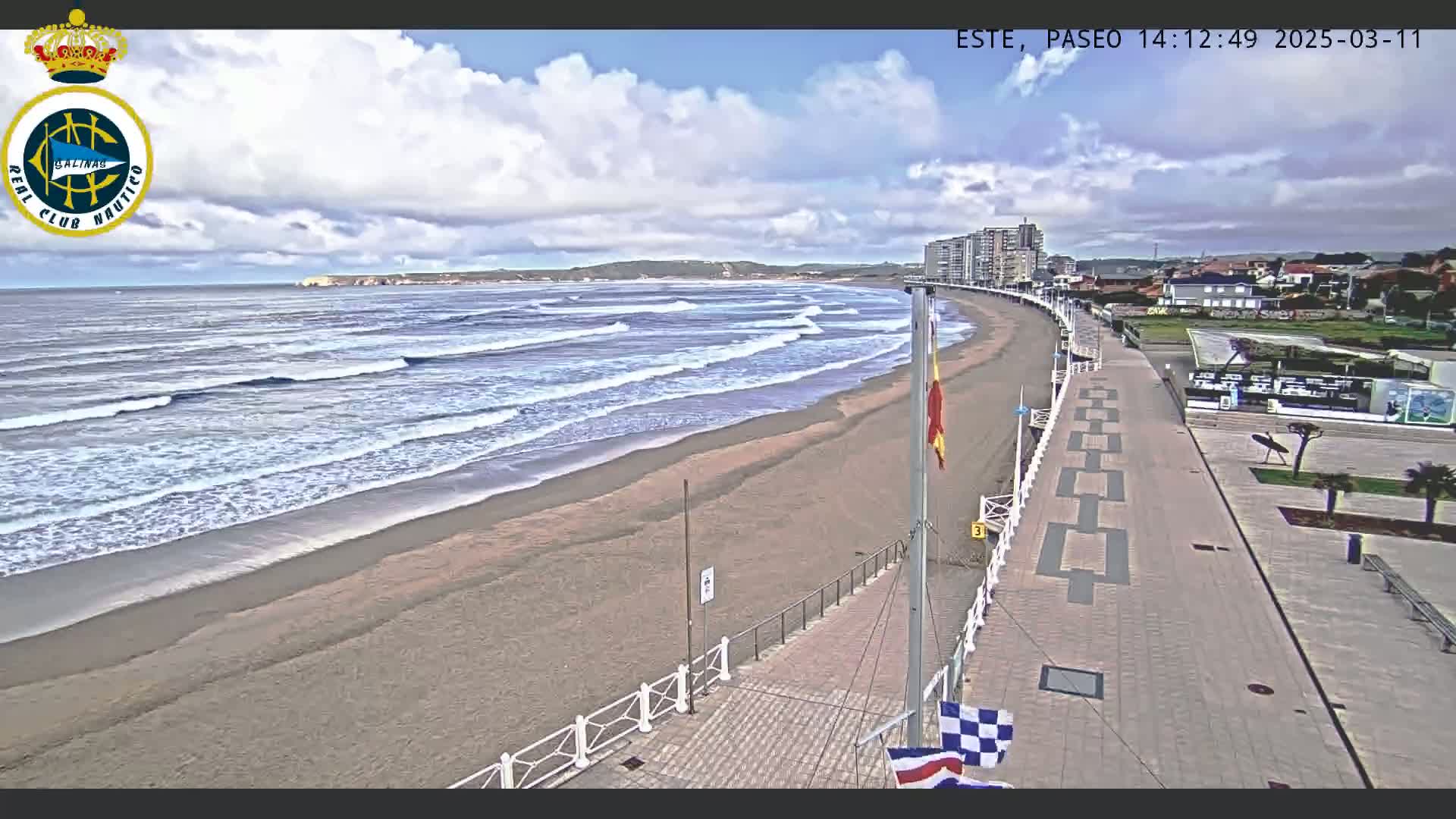 A sandy beach with moderate waves under a partly cloudy sky is bordered by a paved walkway lined with buildings and flags.
