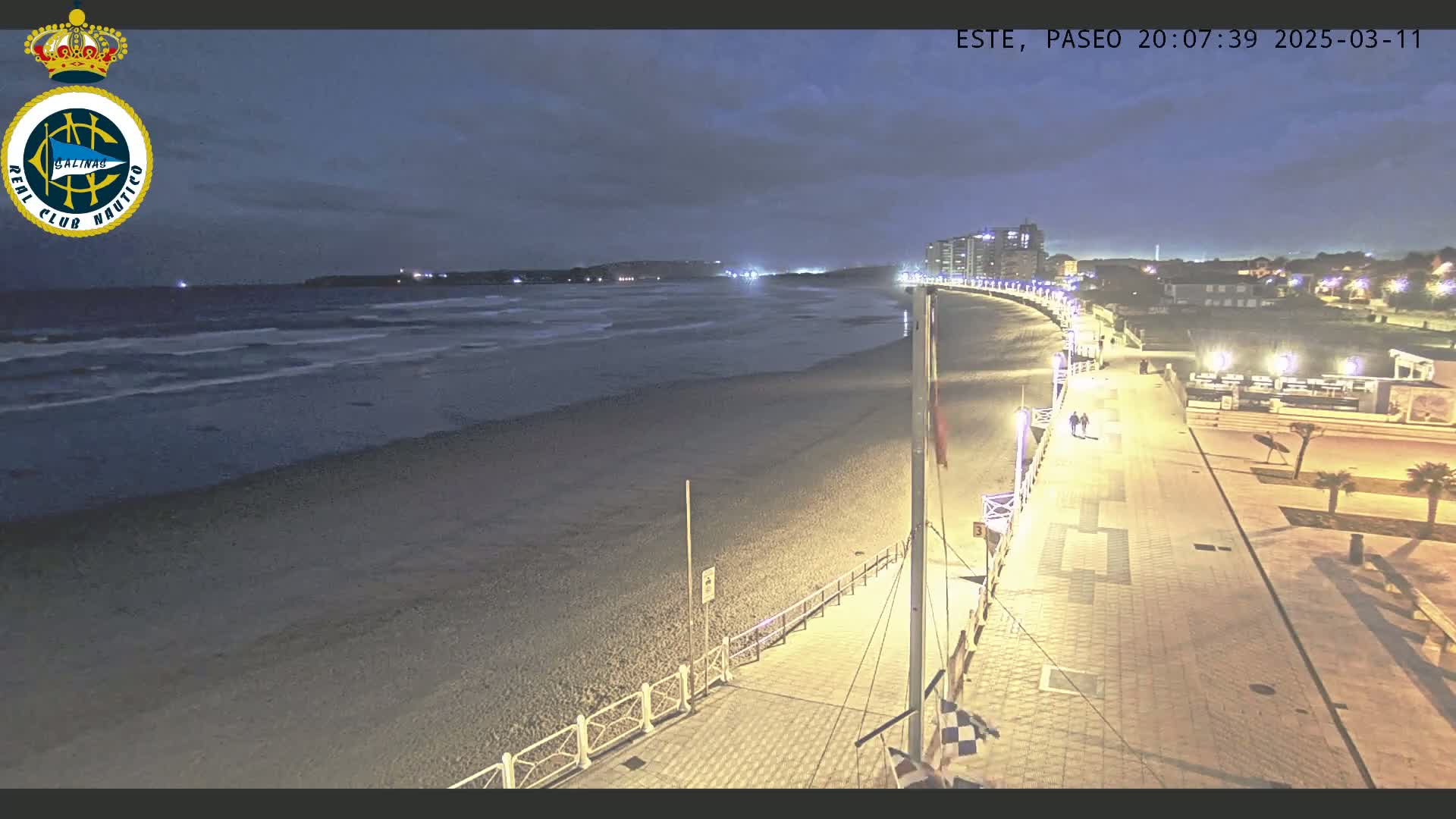 A nighttime view of a beach with a paved walkway lined with lights, leading to buildings in the distance under a mostly cloudy sky.