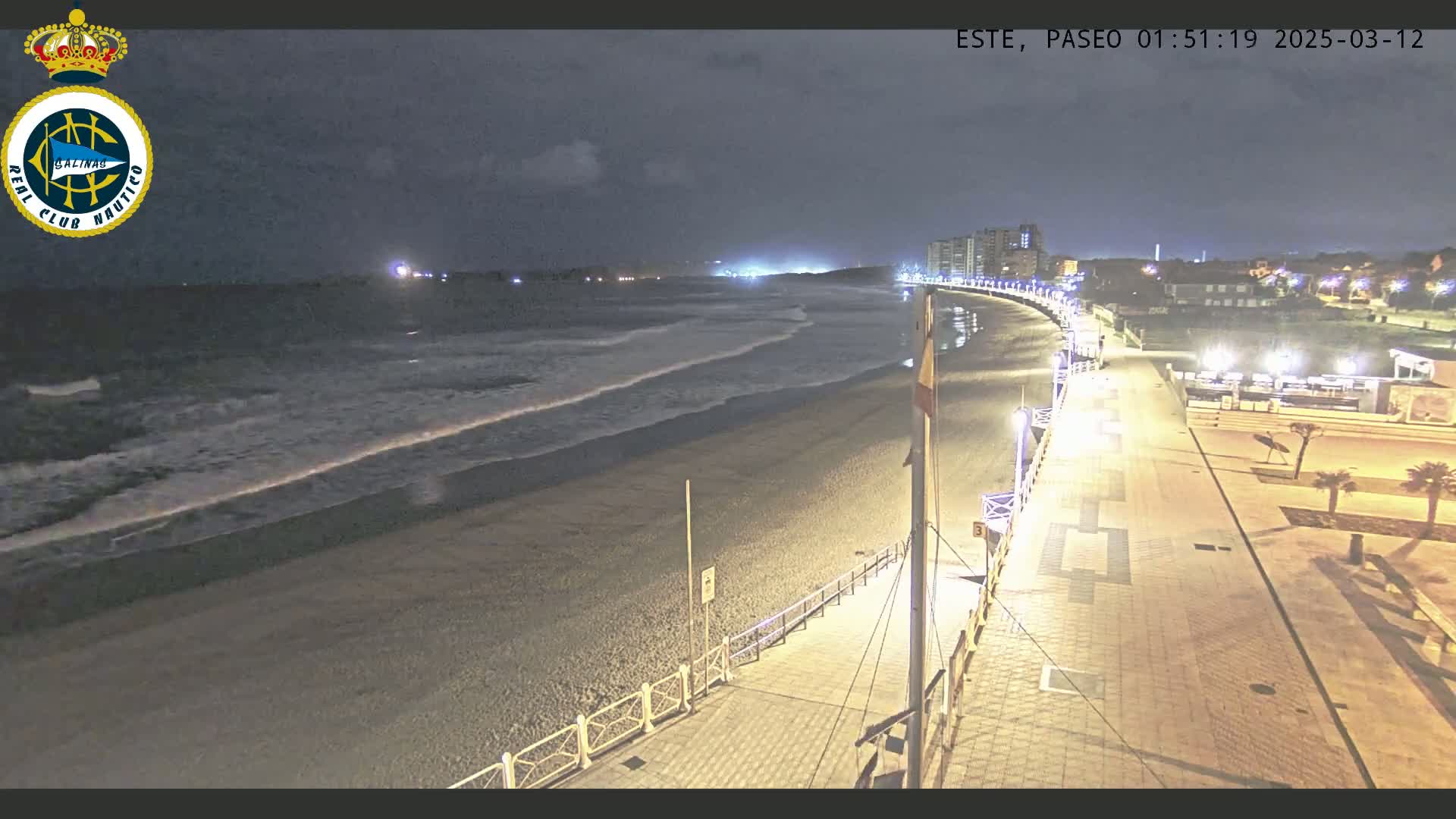 A nighttime view of a beach with gentle waves, a paved boardwalk, and buildings in the distance under a partly cloudy sky.