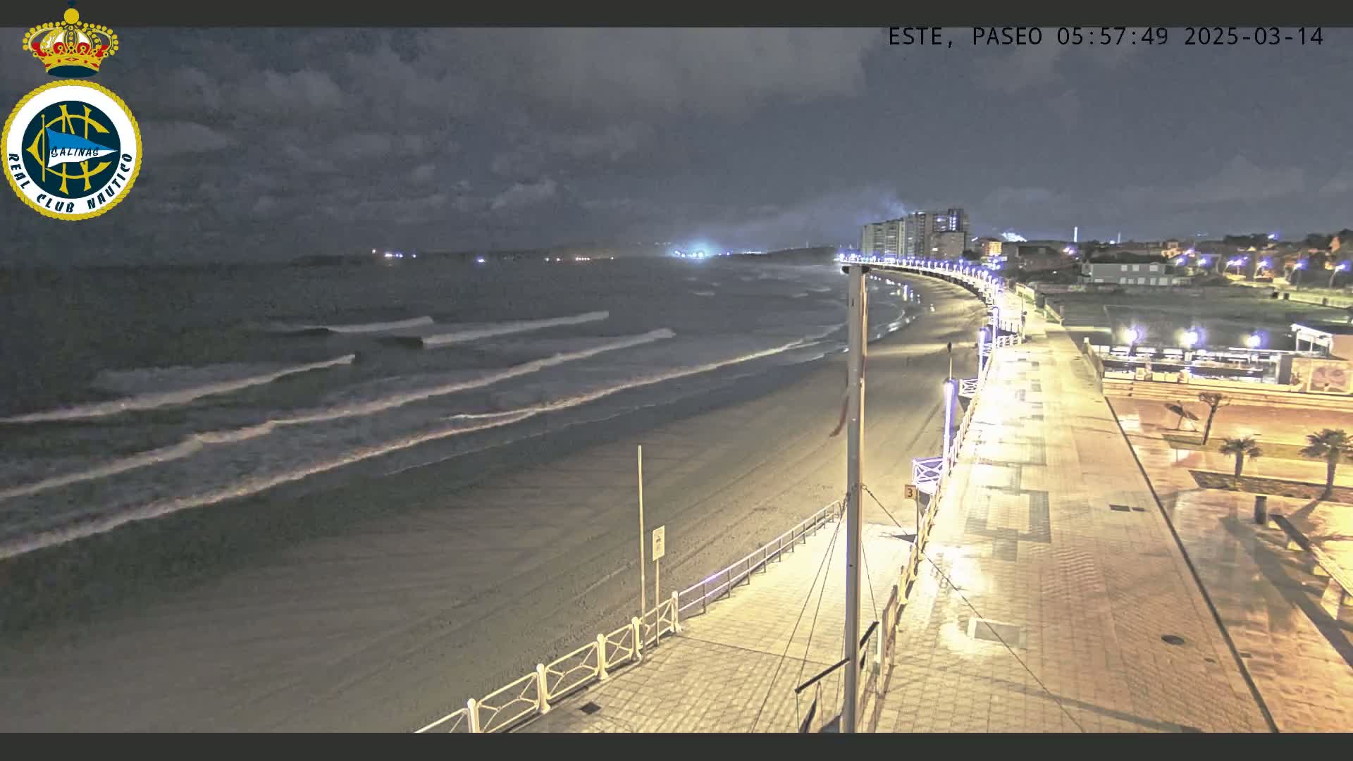 A nighttime view of a beach with gentle waves lapping the shore, a paved walkway lined with lights, and buildings in the background under a cloudy sky.