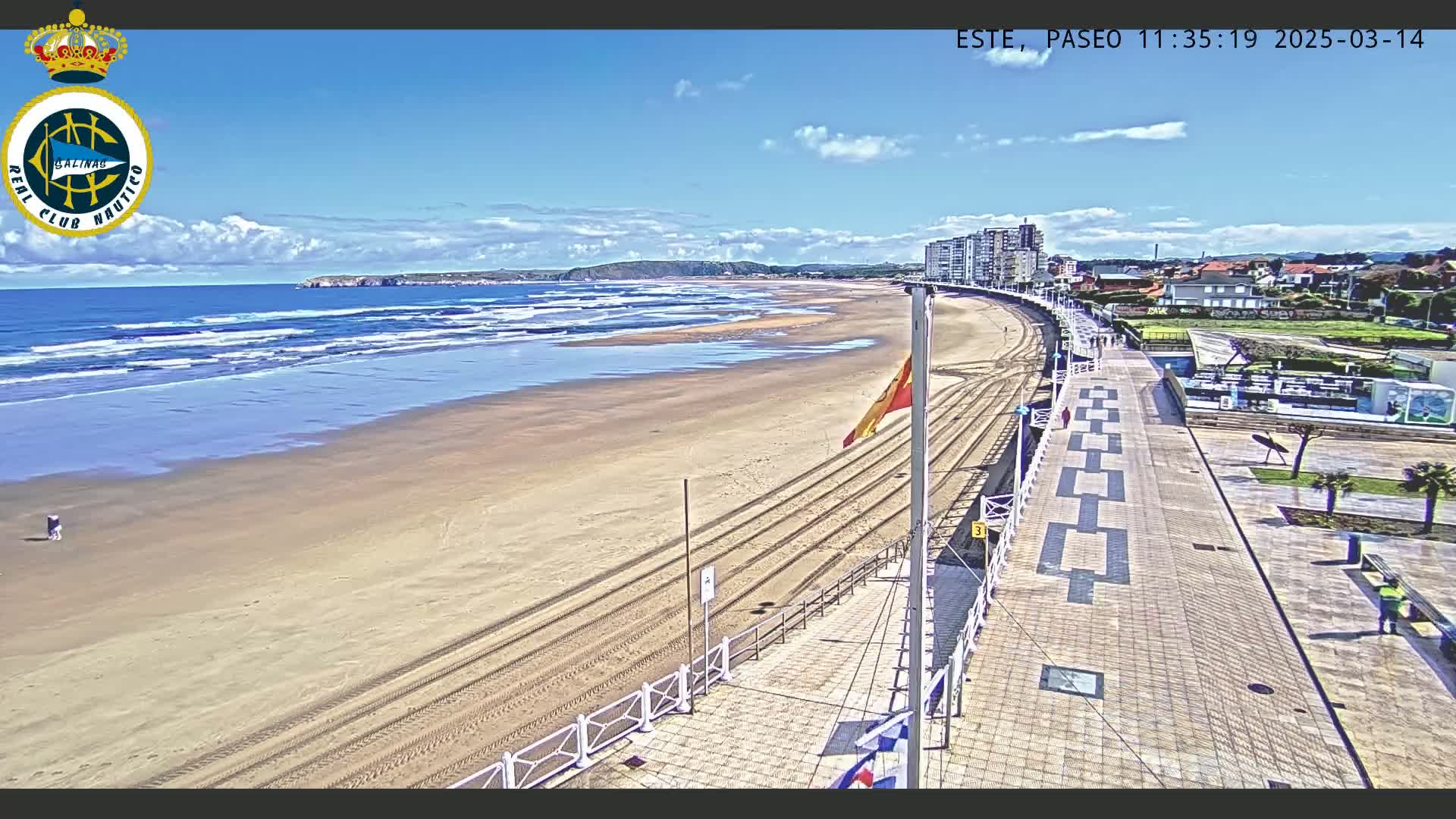 An expansive sandy beach with small waves under a mostly sunny sky is bordered by a paved walkway lined with buildings.