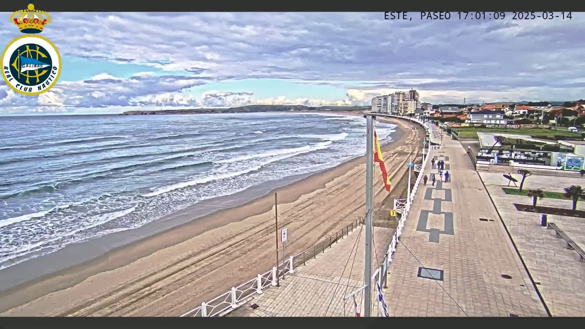 A sandy beach with moderate waves under a partly cloudy sky is next to a paved walkway lined with buildings.