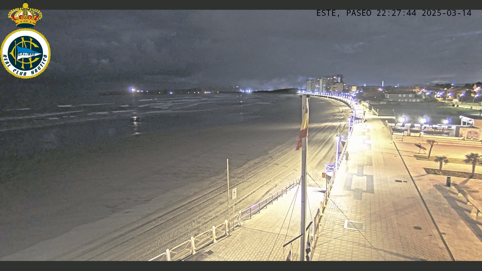 A nighttime view of a mostly empty sandy beach and boardwalk, with calm waves and buildings in the distance under a cloudy sky.