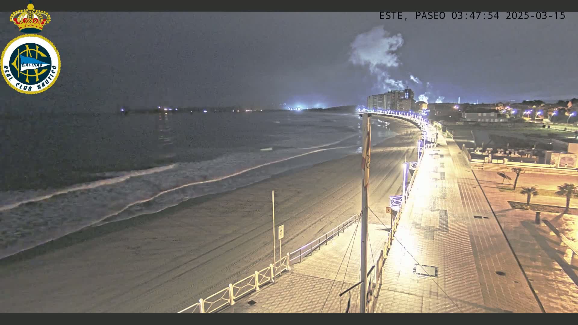 A nighttime view of a mostly empty beach and boardwalk with gentle waves under a dark, cloudy sky.