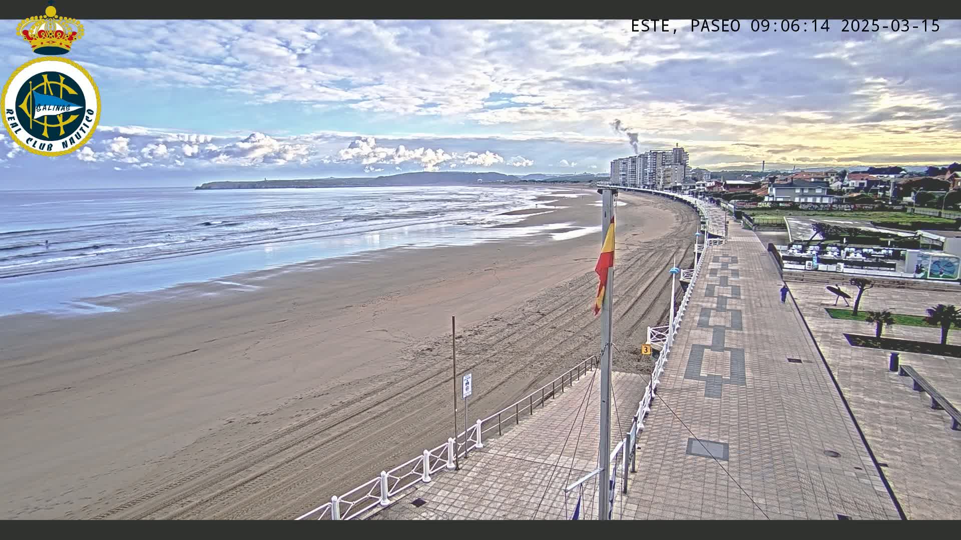 A sandy beach with gentle waves under a partly cloudy sky is seen from above, alongside a paved walkway and buildings in the distance.