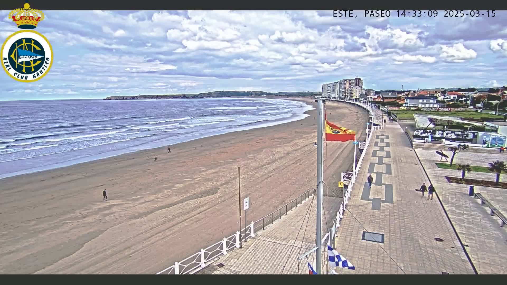 A mostly sunny day at a beach with a sandy shore and gentle waves, viewed from a high vantage point overlooking a paved boardwalk lined with buildings.