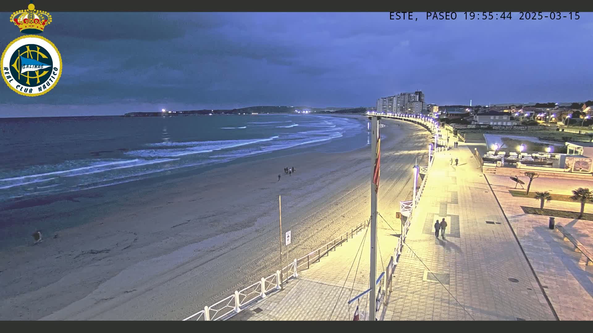 A mostly empty sandy beach at twilight is bordered by a paved walkway with buildings and a few people walking along it, under a cloudy sky.