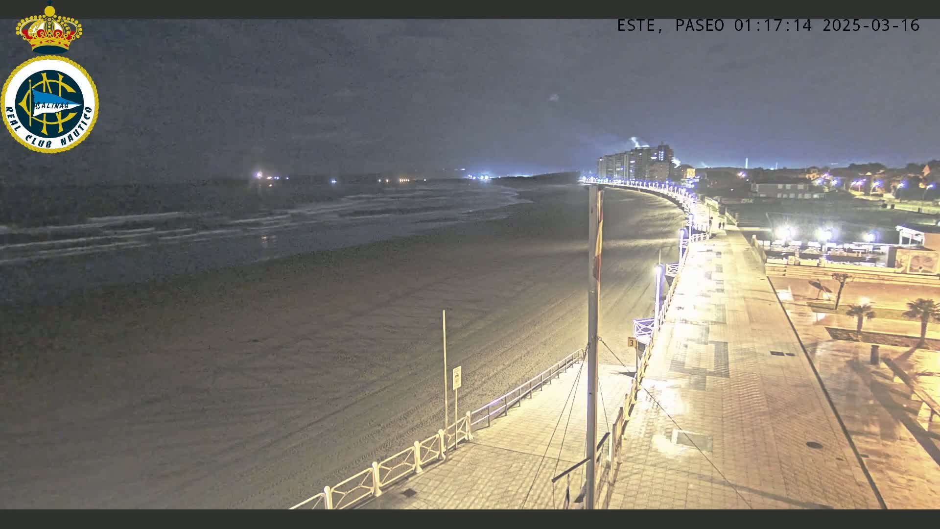 A nighttime view of a mostly empty sandy beach, with a paved boardwalk and buildings visible in the distance under a dark sky.